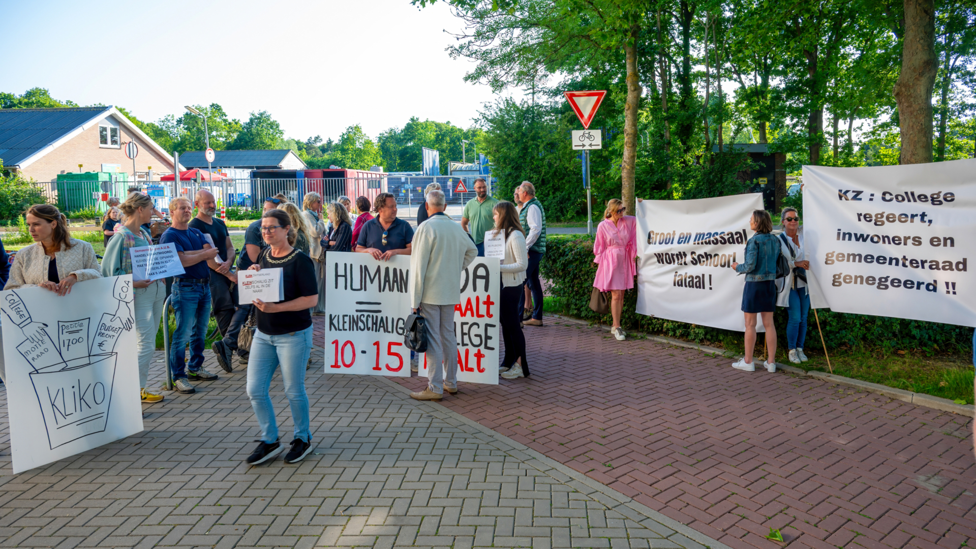 Groep mensen houdt protestborden vast tijdens een demonstratie op een stoep met bomen en gebouwen op de achtergrond.