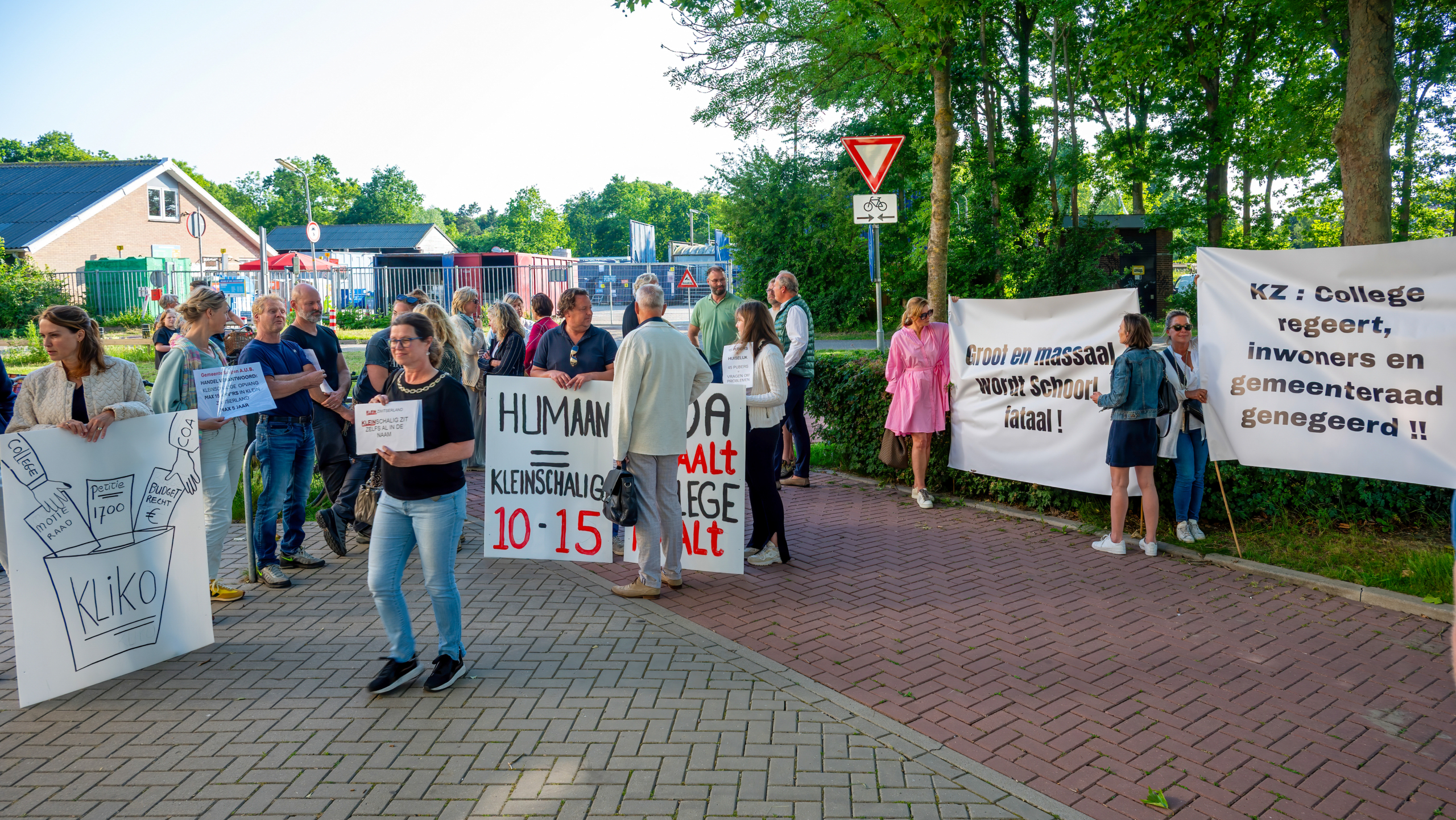 Groep mensen houdt protestborden vast tijdens een demonstratie op een stoep met bomen en gebouwen op de achtergrond.