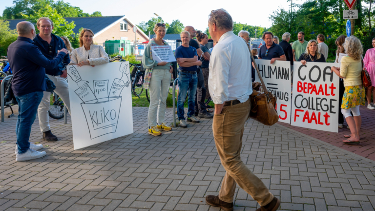 Groep mensen bij een protest met spandoeken die kritische teksten en illustraties tonen, op een trottoir bij een groen omgeving.
