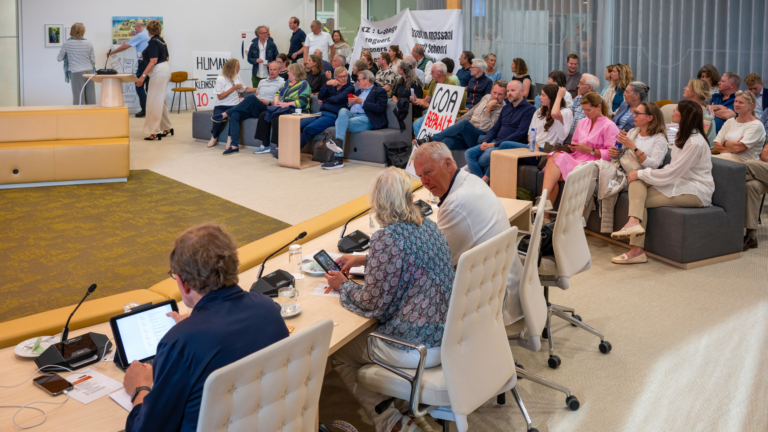 Een gemeentelijke vergaderruimte met diverse mensen die aandachtig luisteren en praten, terwijl sommigen borden vasthouden met teksten. Op de voorgrond zitten mensen aan een tafel met microfoons en papier, terwijl op de achtergrond een groep mensen op banken zit.