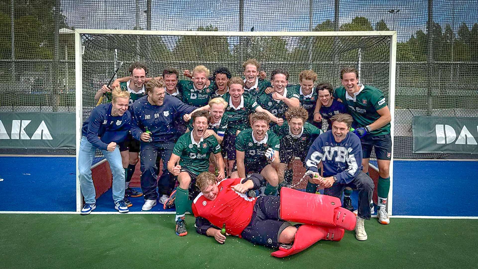 Een mannenhockeyteam viert enthousiast met een lach, omringd door een doelnet op een hockeyveld, met een doelman in rood liggend vooraan, en enkele spelers met waterflessen.