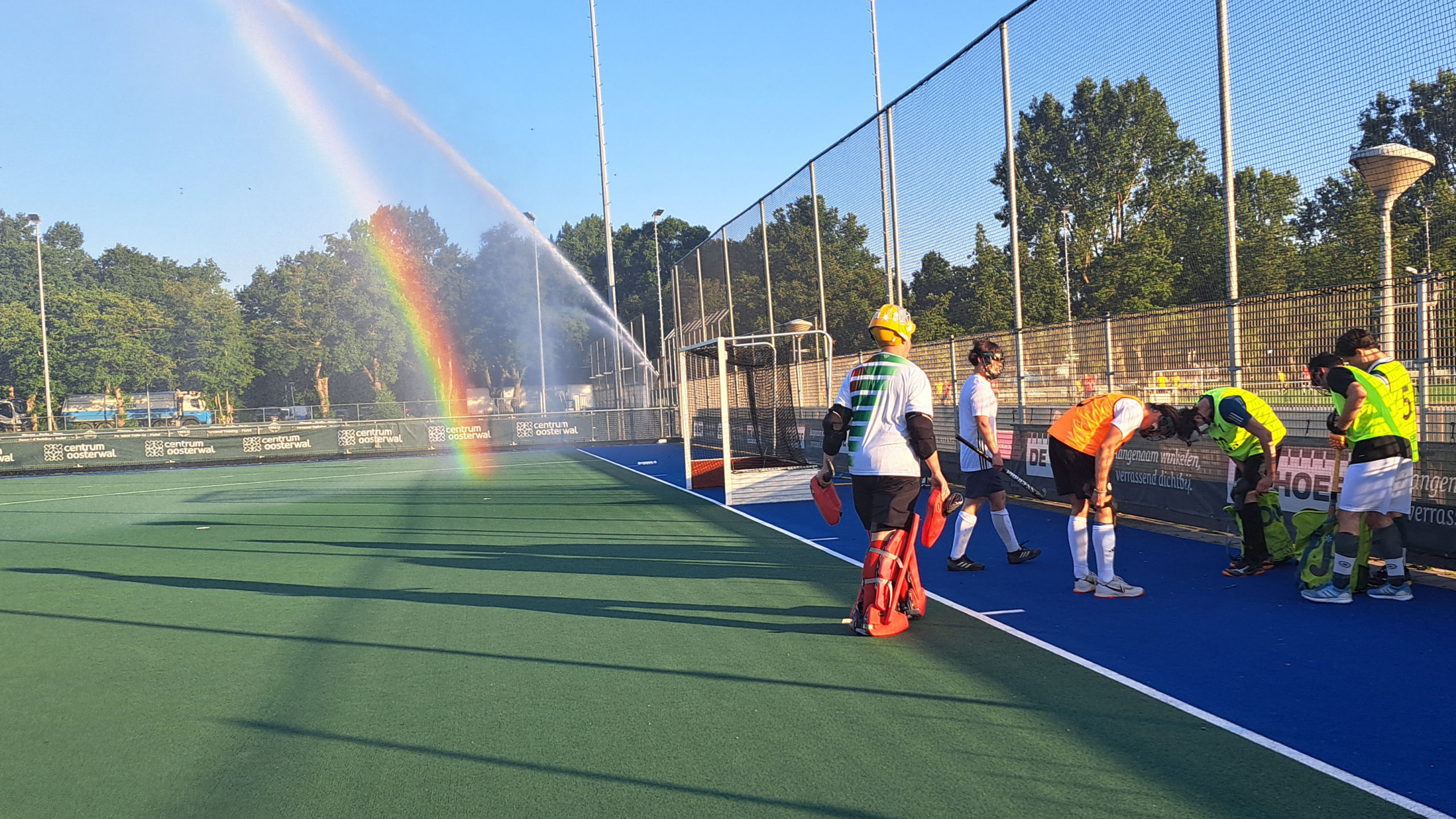 Hockeyspelers op een veld, met een regenboog zichtbaar door een sproeiende sprinkler op de achtergrond.