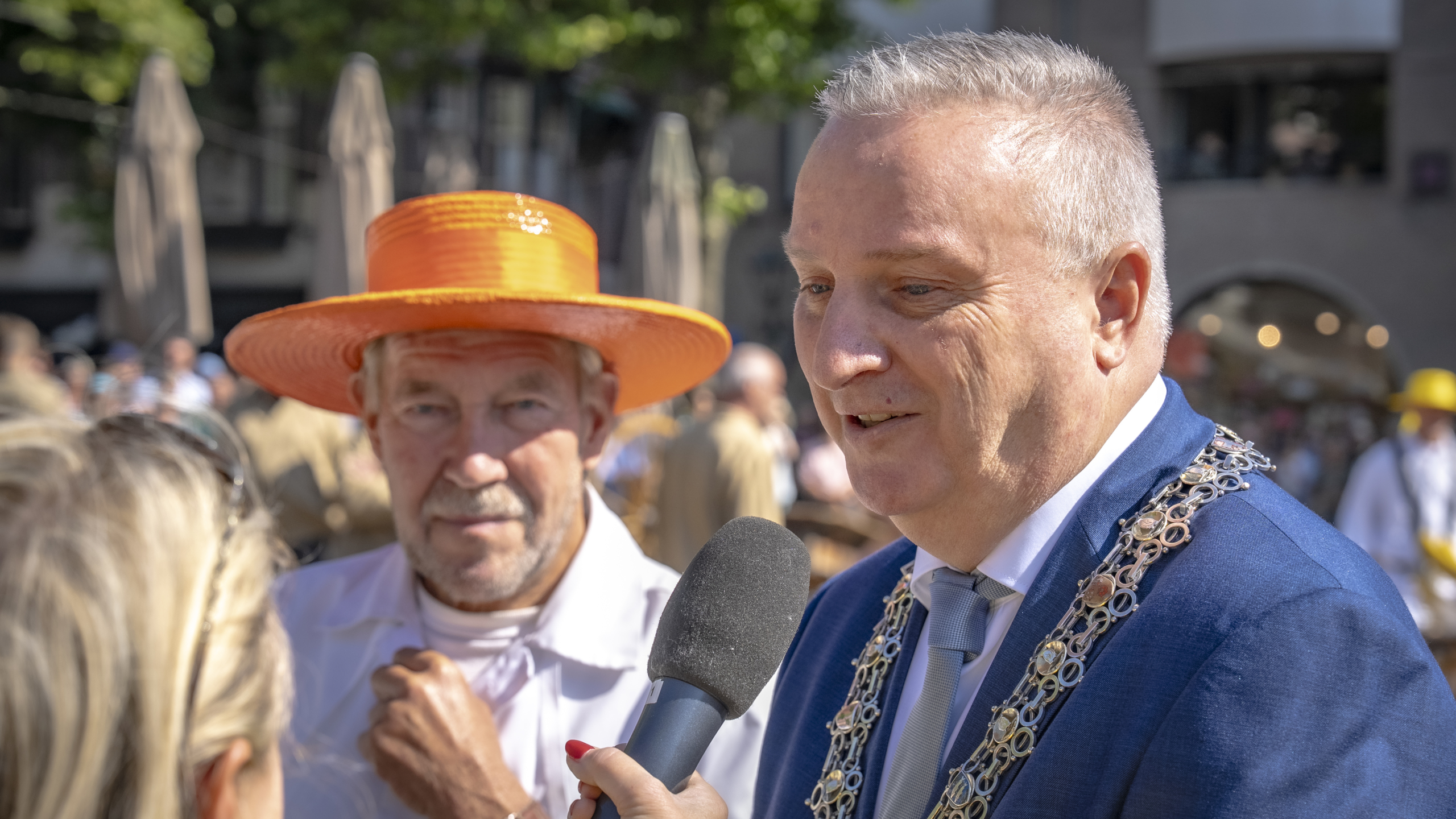 Een man in een blauw pak met een zilveren ketting wordt geïnterviewd met een microfoon, terwijl er een andere man met een oranje hoed naast hem staat in een drukke buitenomgeving.