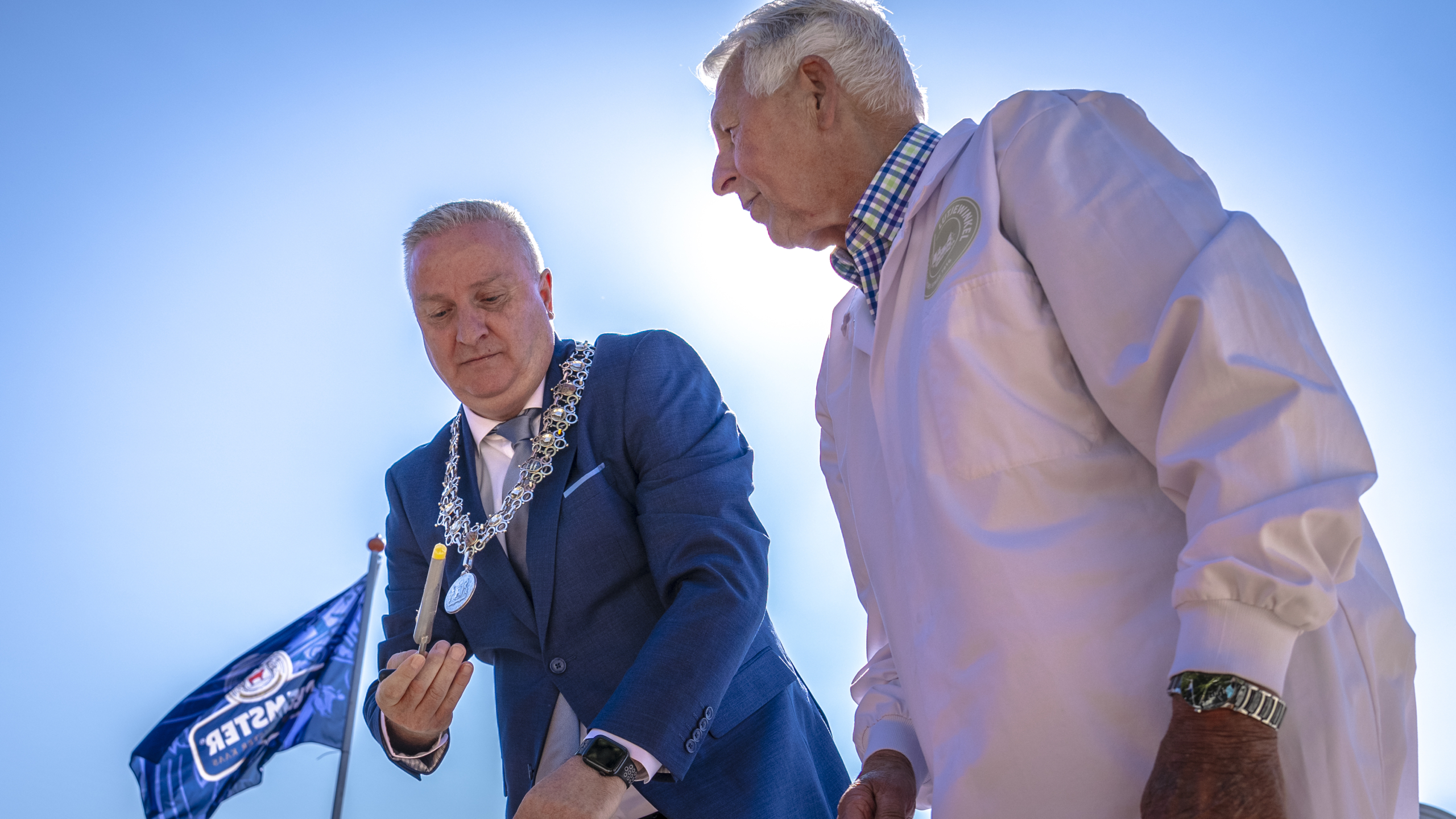 Twee mannen, een in een blauw pak en de ander in een witte jas, bekijken een object in een heldere omgeving met een blauwe lucht en een vlag in de achtergrond.