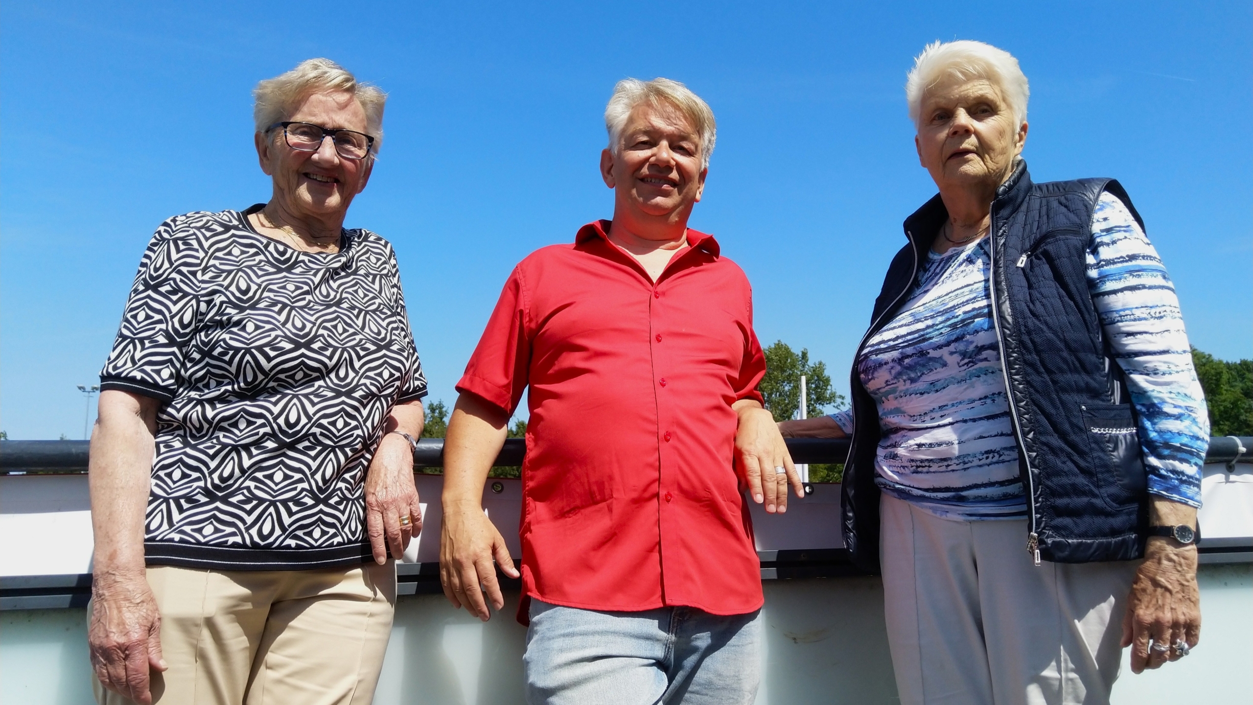 Drie oudere volwassenen staan samen buiten tegen een leuning, onder een heldere blauwe lucht.