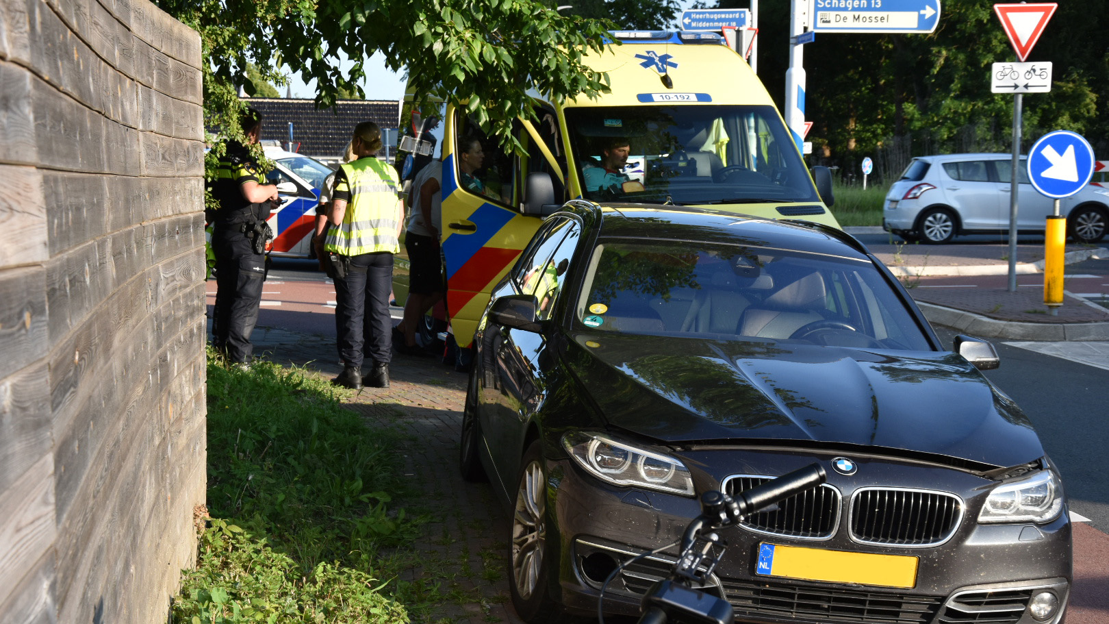 Een politieagent staat naast een politieauto en ambulance bij een verkeersongeval met een zwarte BMW op een rotonde.