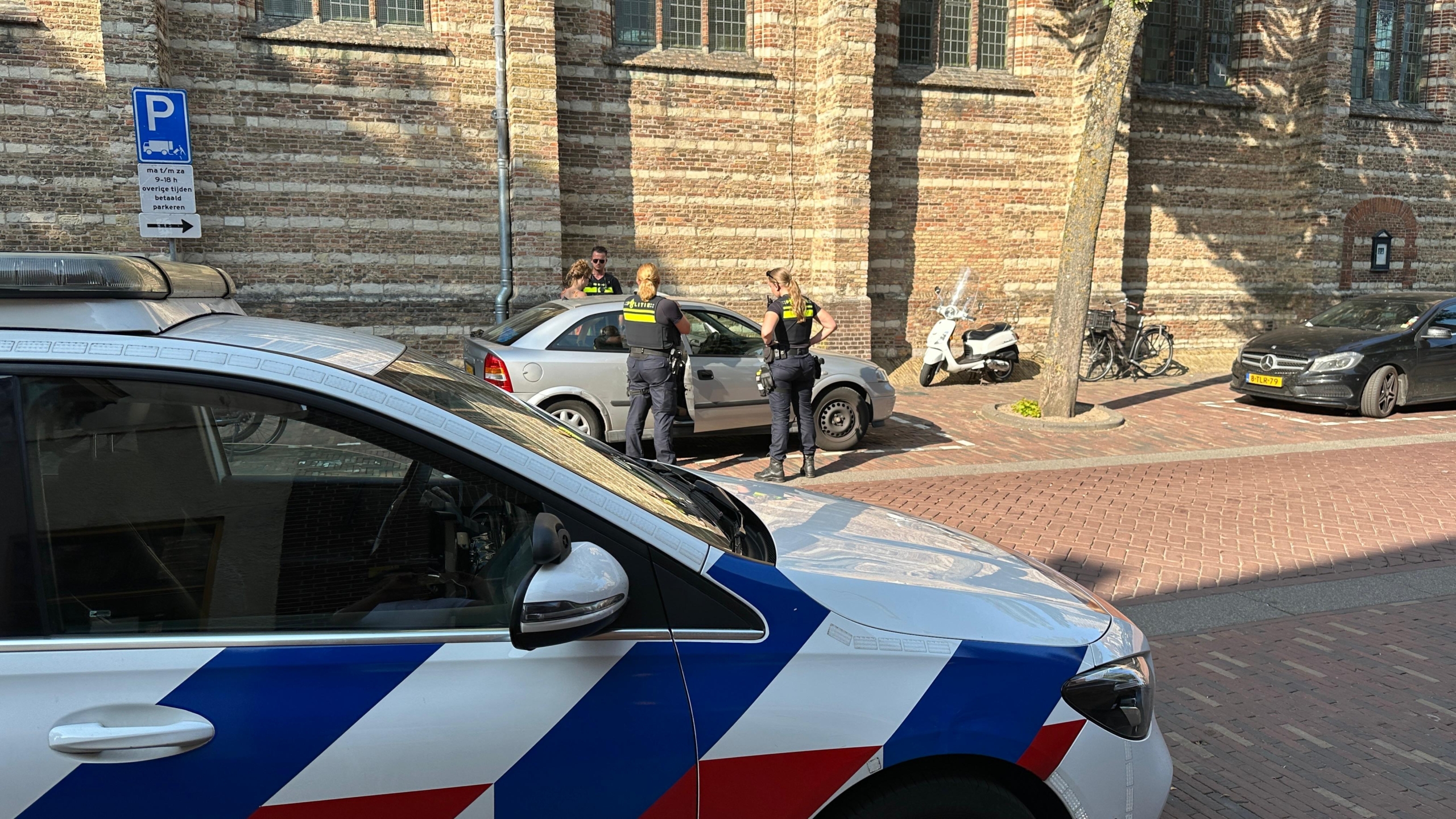 Politieagenten staan naast een geparkeerde auto en praten met een persoon op straat; op de voorgrond is een politieauto zichtbaar.