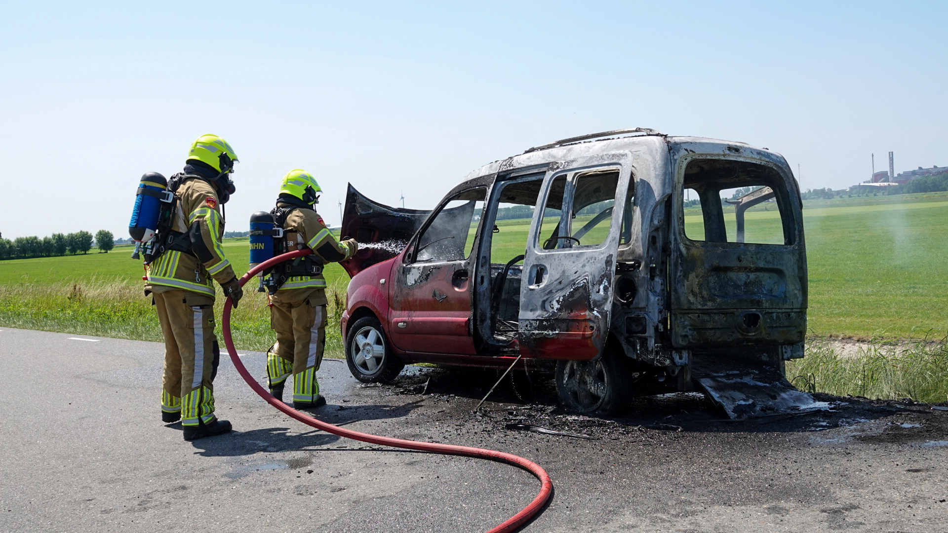 Twee brandweerlieden in beschermende kleding blussen een uitgebrande auto langs een weg met behulp van een brandslang, tegen een achtergrond van groene velden en een blauwe lucht.
