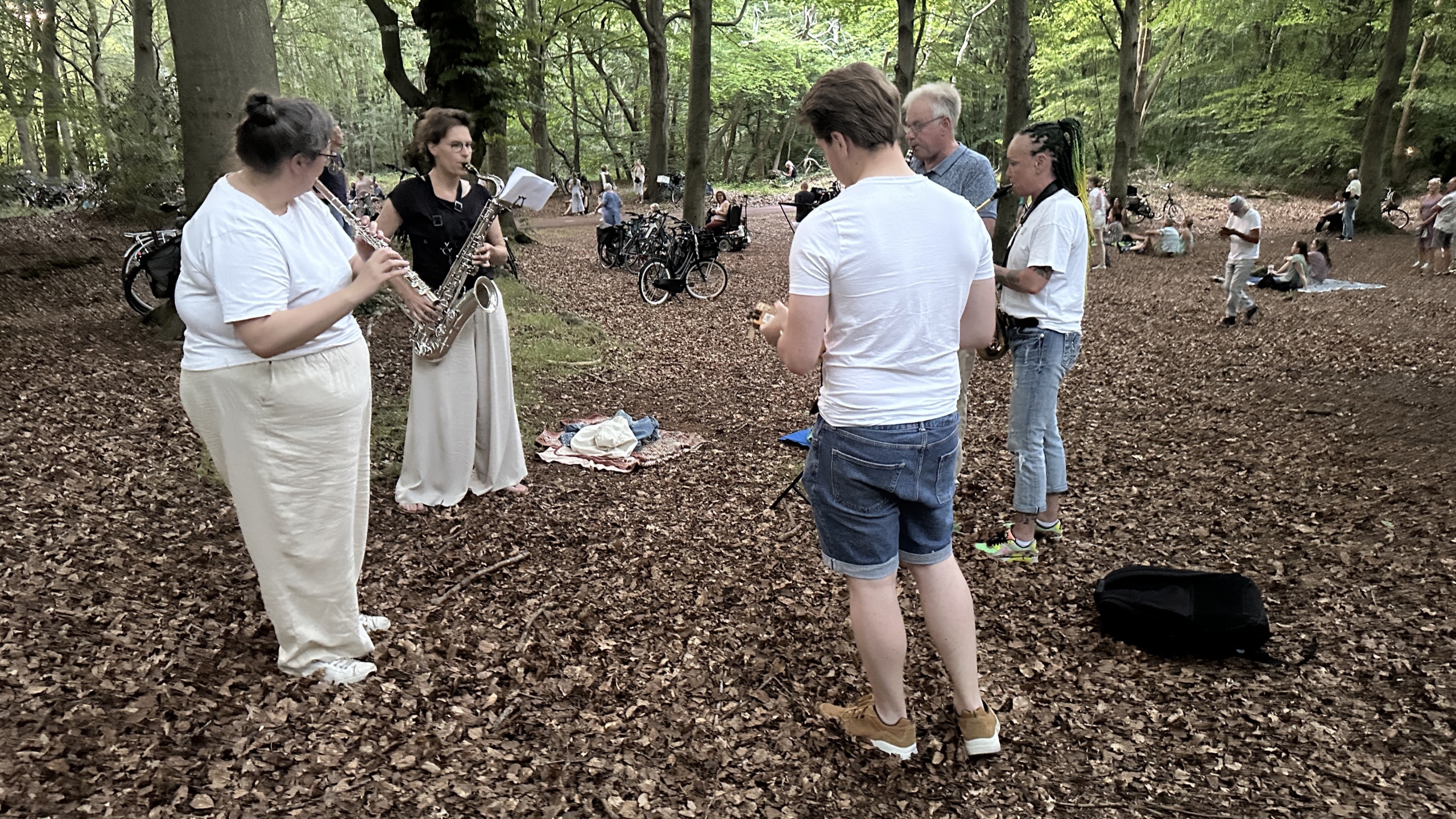 Een groep mensen speelt muziek in een bos, met een vrouw die een saxofoon bespeelt en een andere die een dwarsfluit bespeelt, omringd door anderen die aandachtig kijken. De bosgrond is bedekt met bladeren en er staan enkele fietsen in de achtergrond.