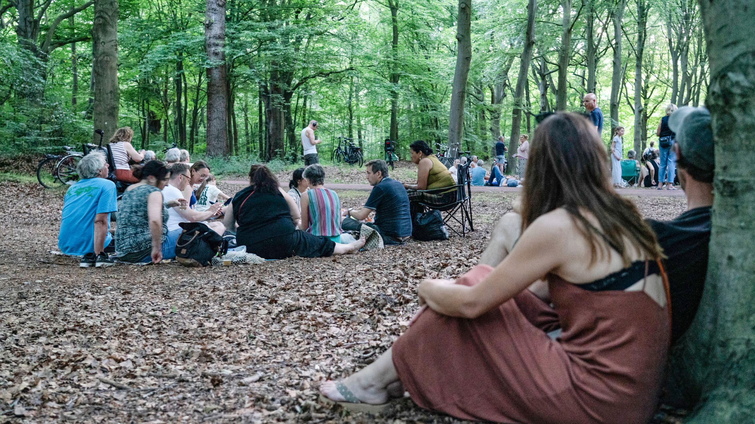 Mensen zitten in een bosrijke omgeving, sommigen op stoelen en anderen op de grond, terwijl ze ontspannen en met elkaar praten.