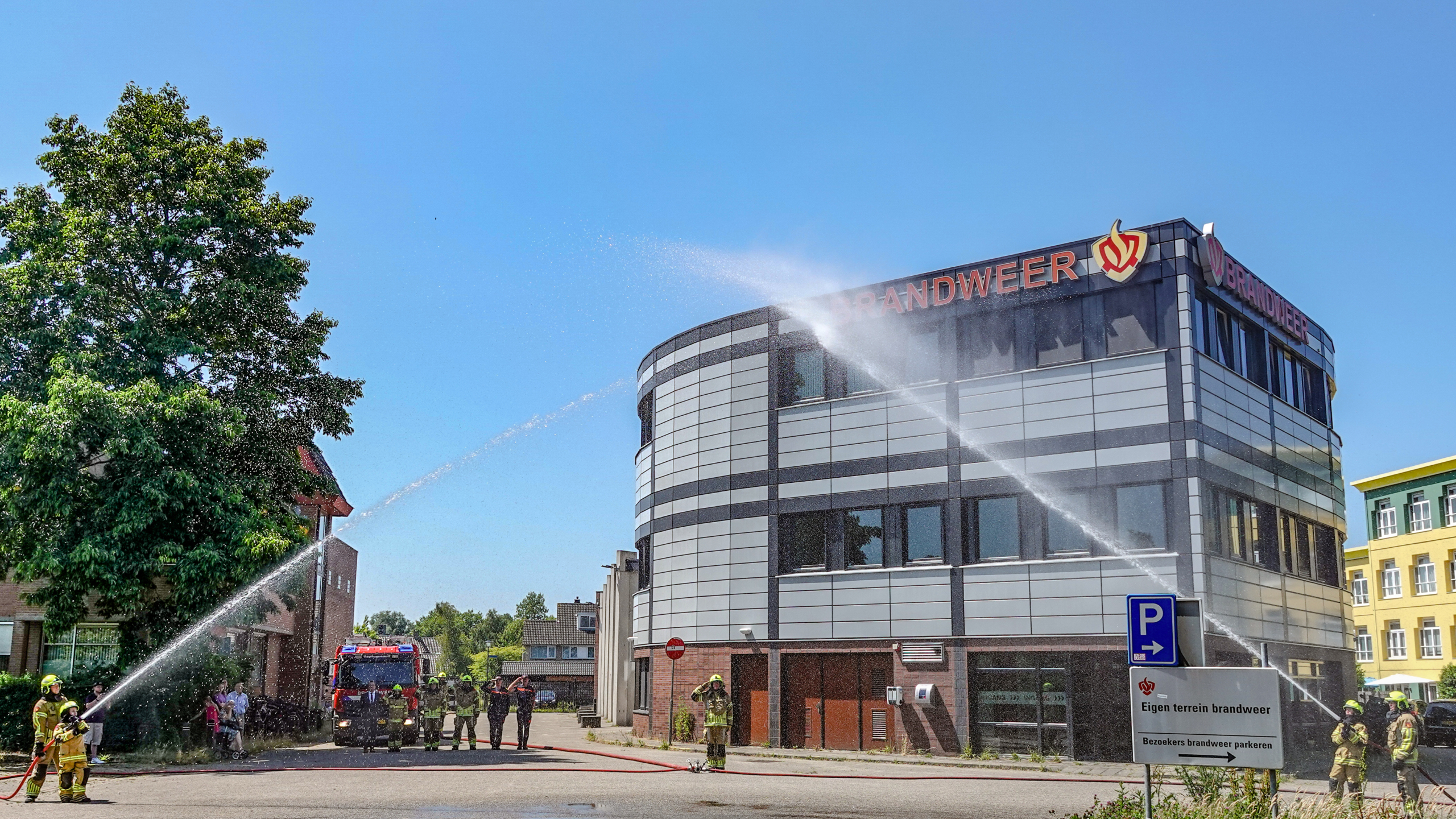 Brandweermannen spuiten water met brandslangen op een gebouw met het bord 'Brandweer', terwijl een brandweerwagen op de achtergrond staat en toeschouwers toekijken.