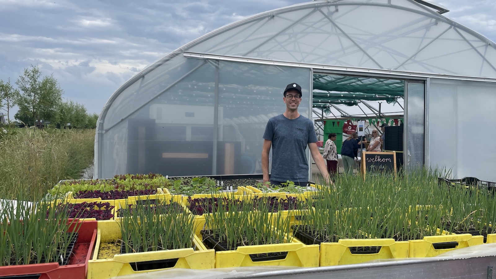 Een man staat voor een kas met bakken met jonge planten en scheuten ervoor, terwijl meerdere mensen binnen de kas praten. Er hangt een bord met 'welkom' bij de ingang. De lucht is bewolkt.