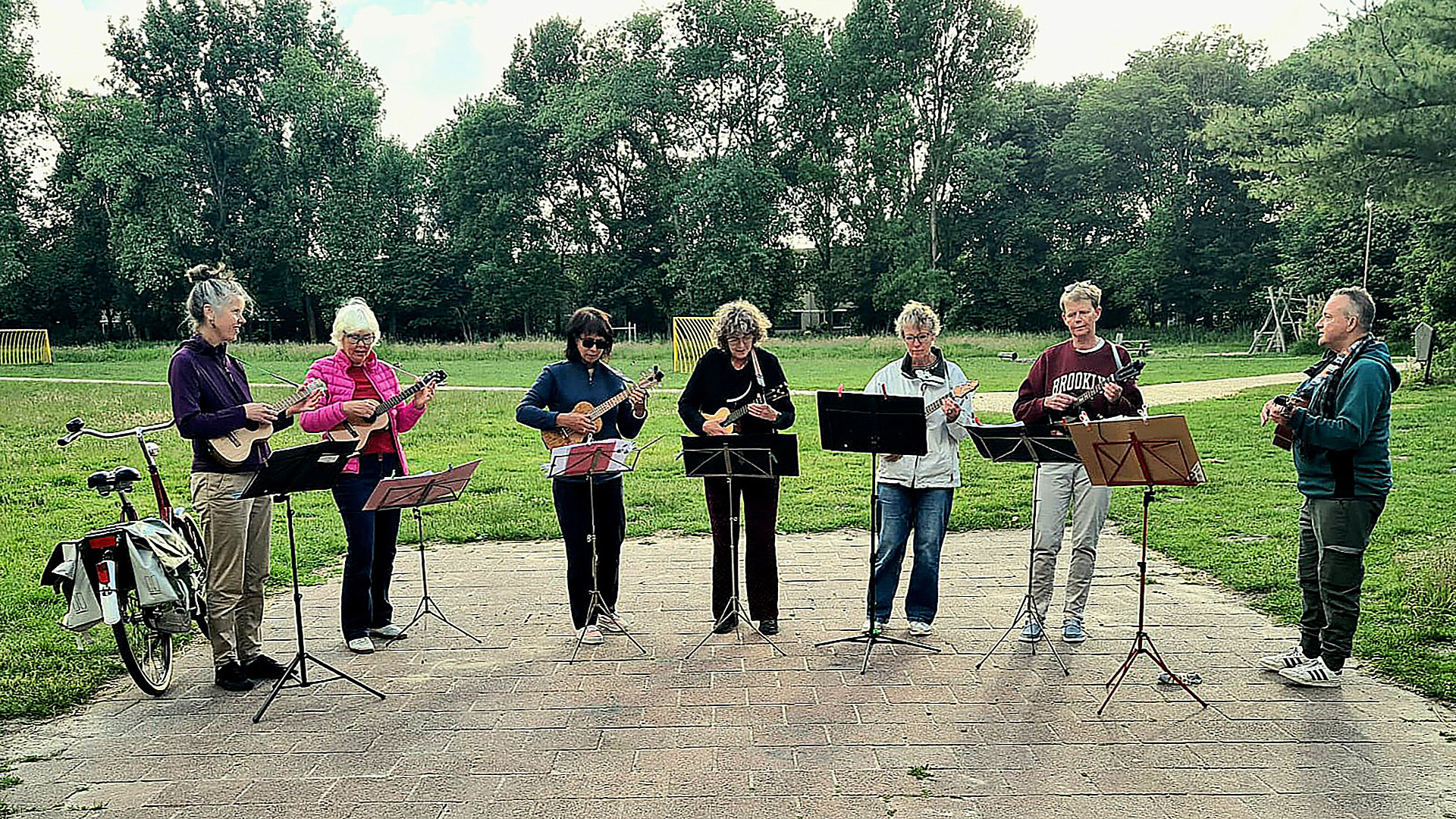 Groep mensen speelt muziek op ukuleles in een park, met muziekstandaards voor zich en bomen op de achtergrond.