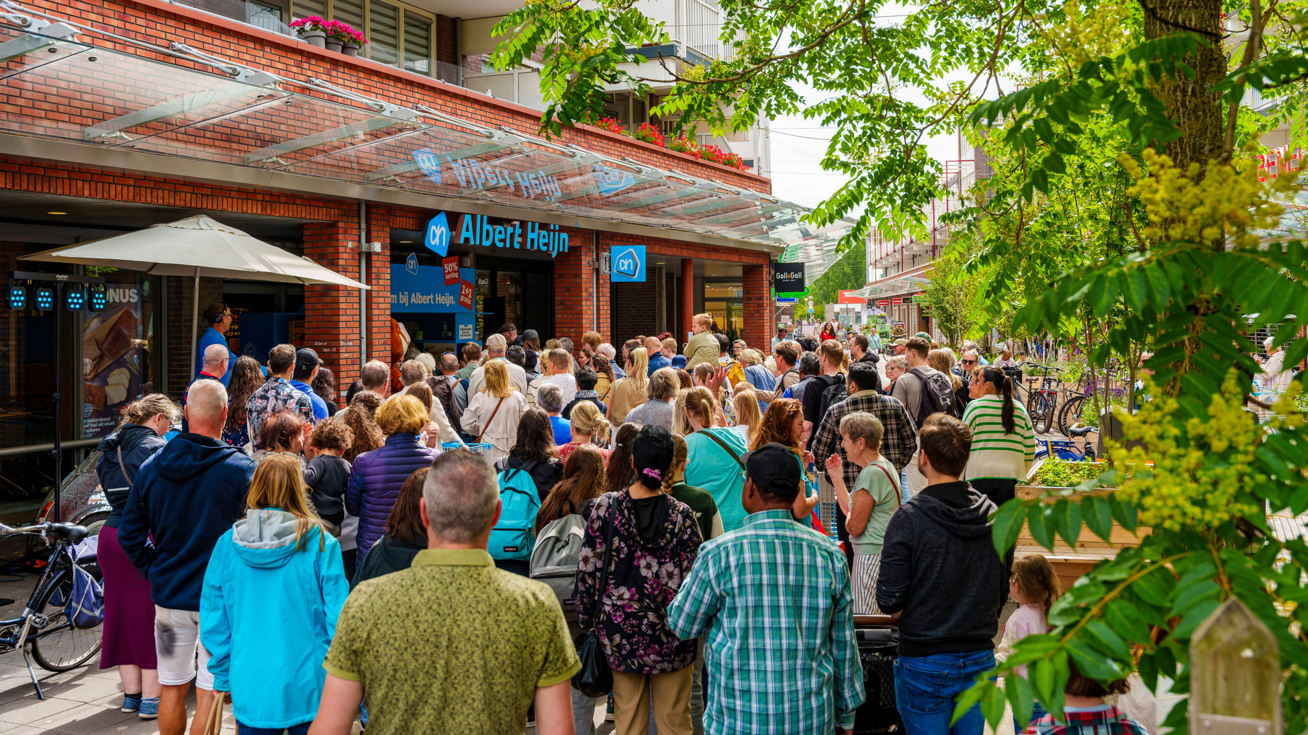 Een grote groep mensen staat in de rij voor de ingang van een Albert Heijn supermarkt, omringd door groene bomen en fietsen.