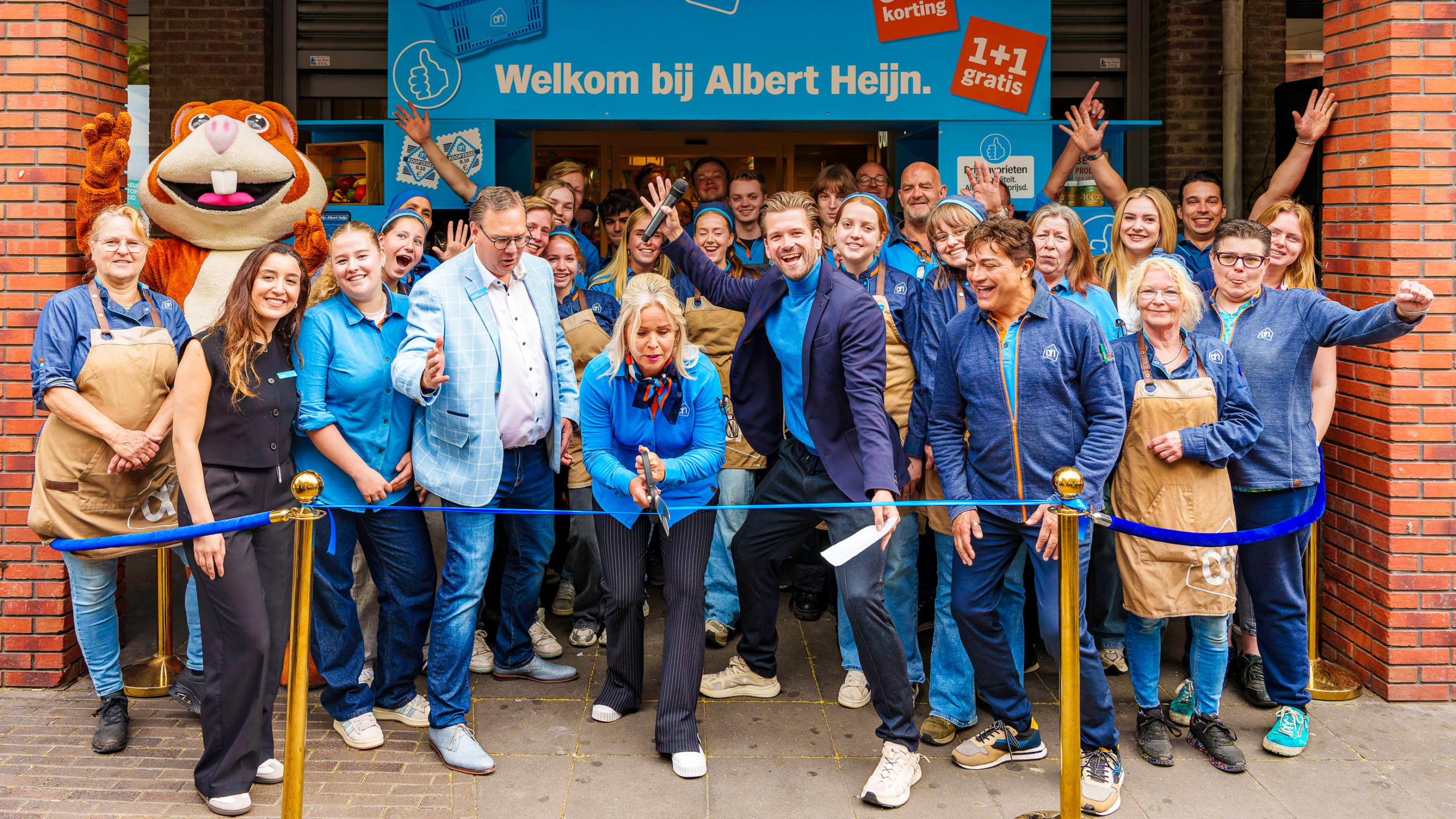 Een groep mensen voor een Albert Heijn-supermarkt poseert bij een lint voor een openingsevenement. Een vrouw knipt het lint door, omringd door medewerk(st)ers in blauwe kleding en een mascotte in een dierenkostuum. De sfeer is feestelijk en vrolijk.