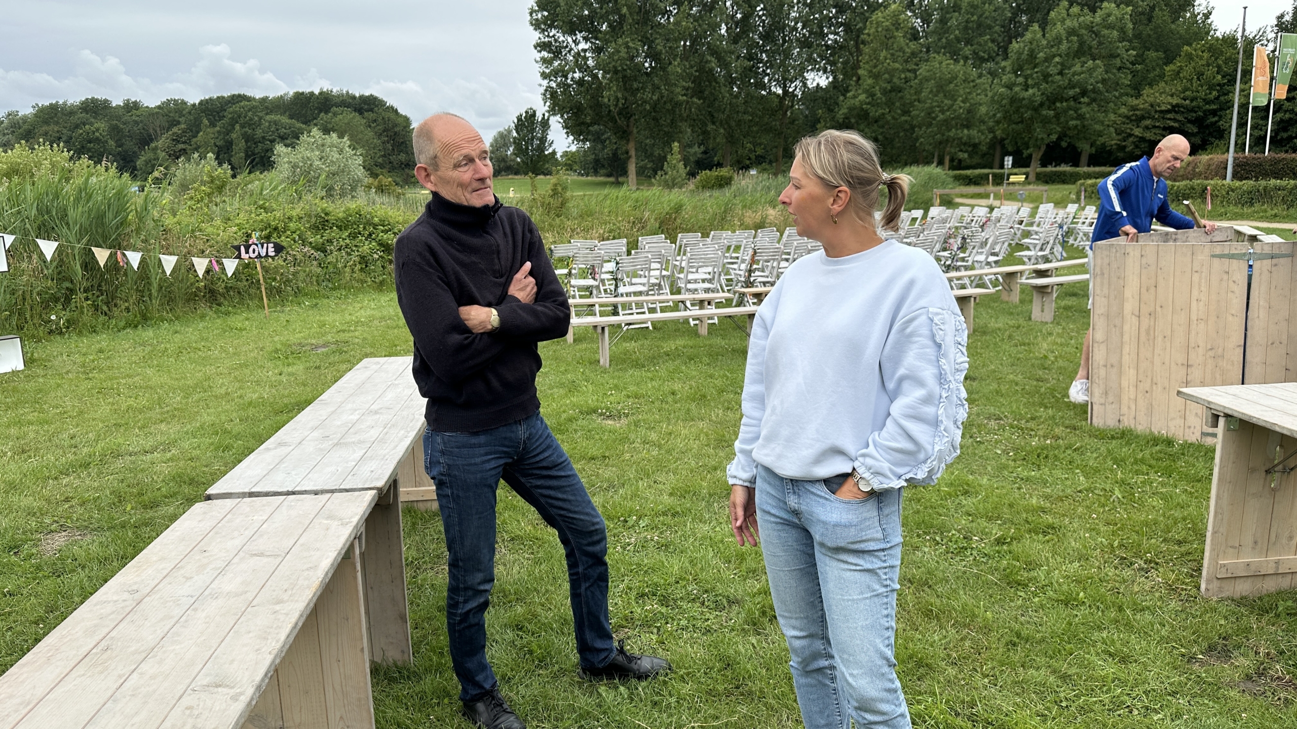 Twee personen staan op een grasveld en praten met elkaar, omringd door stoelen en vlaggetjes met de tekst 'LOVE'.