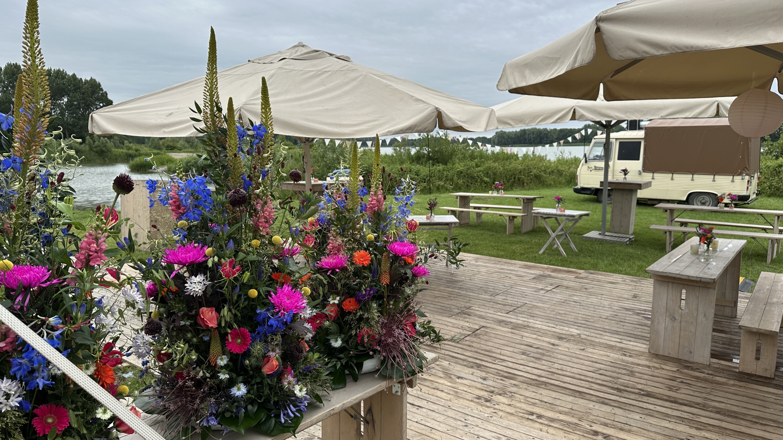 Buitenruimte met houten vlonder, kleurrijke bloemstukken, parasols en een vintage bestelwagen op een grasveld met op de achtergrond een rivier en bomen.