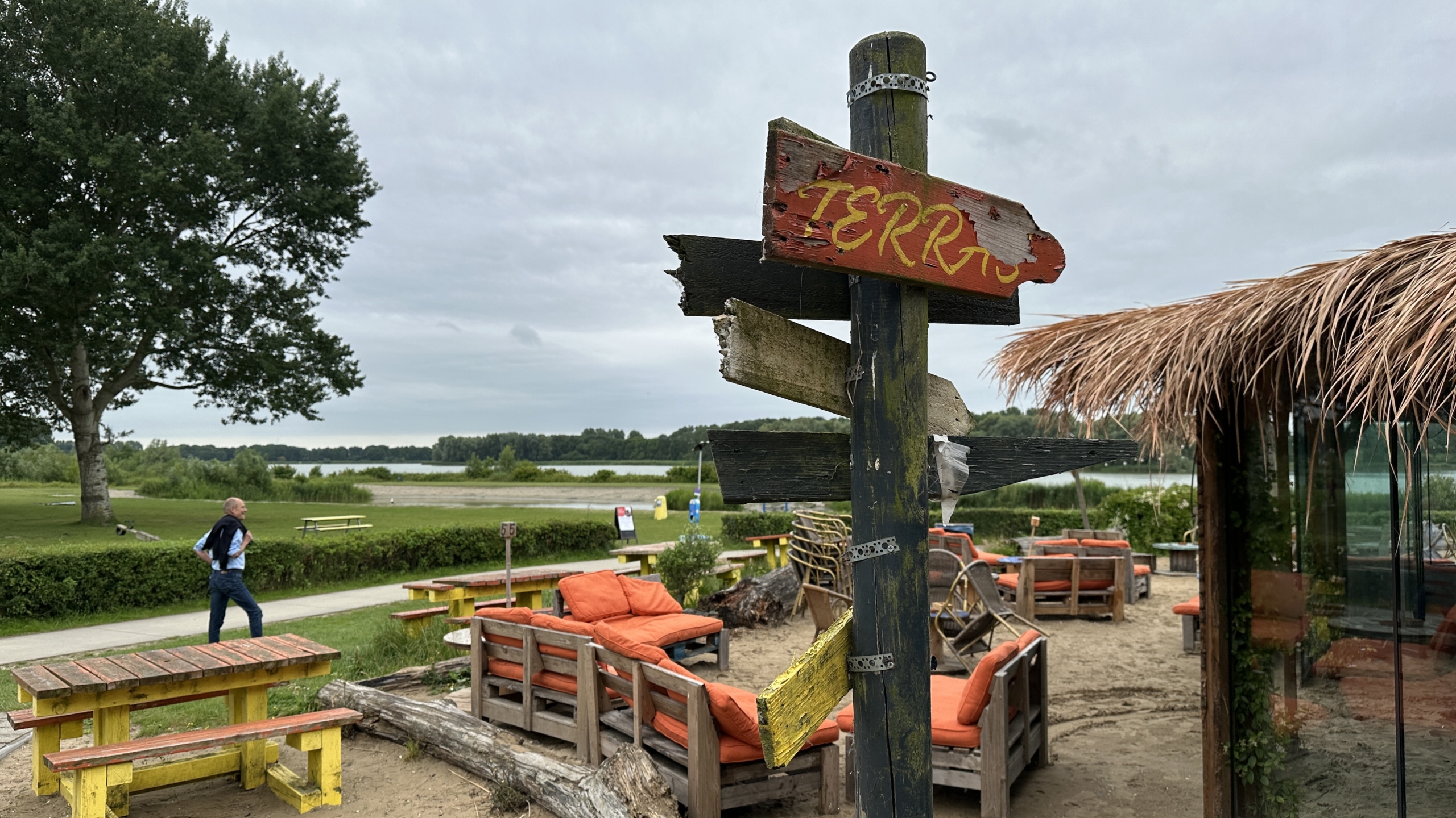 Een rustieke houten wegwijzer met de tekst 'TERRAS' wijst naar een strandterras met kleurrijke meubels en een rieten dak, gelegen aan een rivier met groen landschap op de achtergrond.