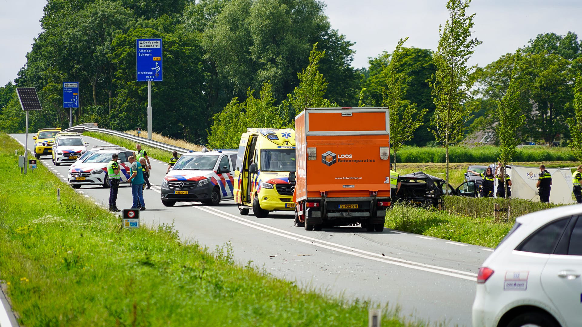 Verkeersongeval op een landelijke weg met politie, ambulancevoertuigen en een bergingsvoertuig aanwezig. Een oranje vrachtwagen blokkeert gedeeltelijk de weg, terwijl schade zichtbaar is aan een zwart voertuig aan de rechterkant. Hulpverleners staan op de weg en in de berm naast een bord richting Heerhugowaard.
