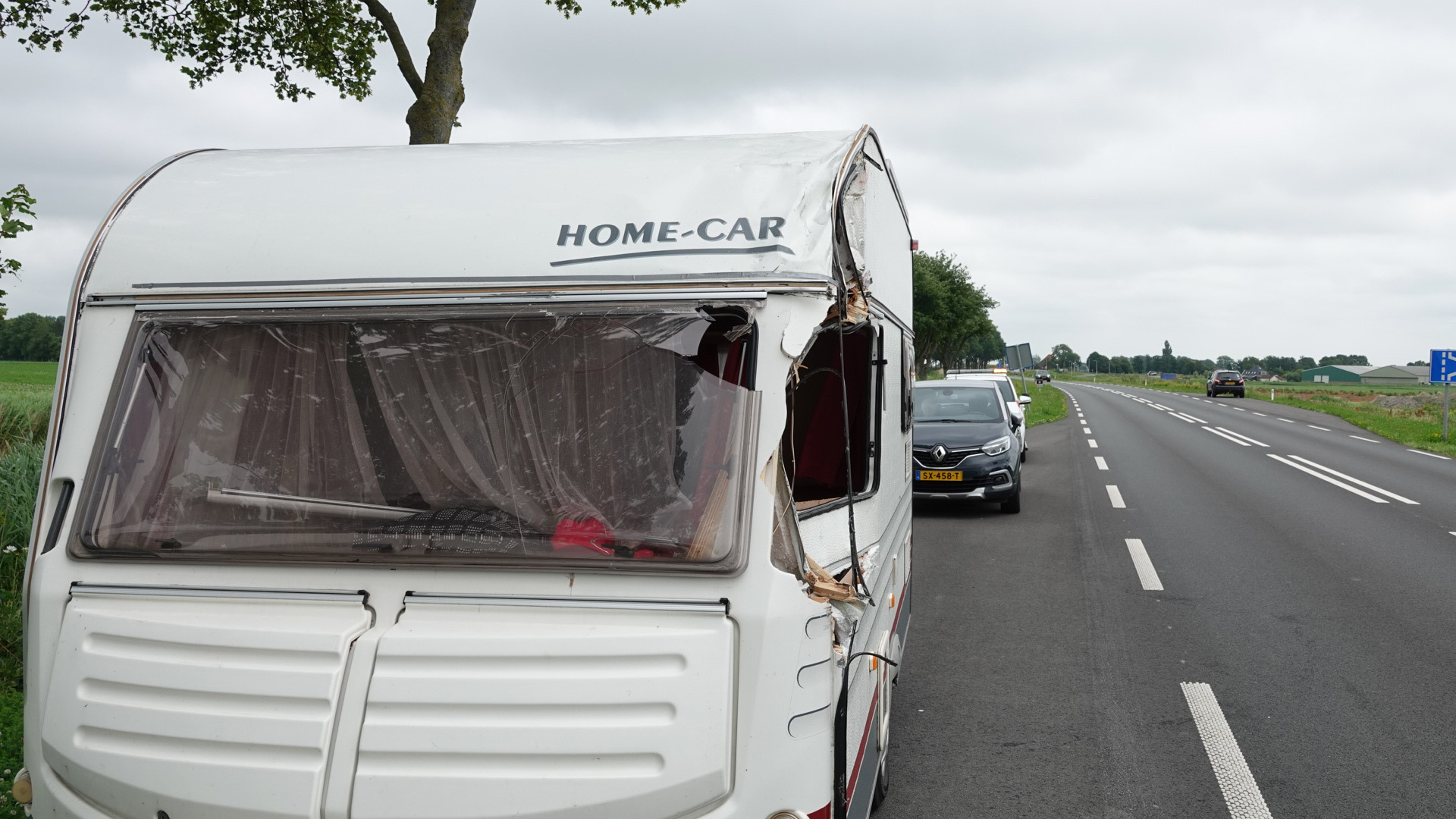 Beschadigde caravan met een kapot raam en het merk 'HOME-CAR', geparkeerd langs een landweg met een gezonken locatie aan de kant.