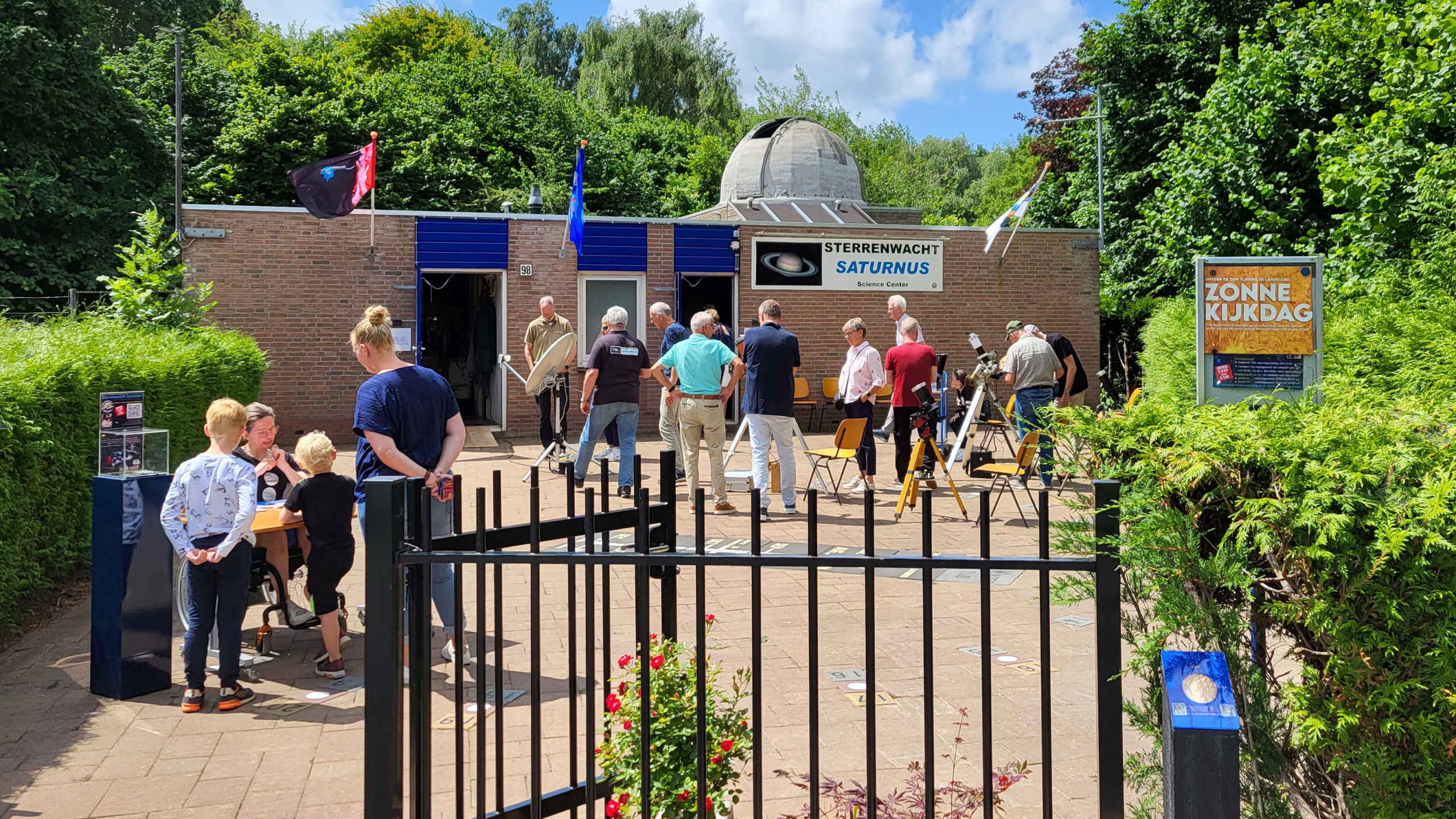 Mensen verzamelen zich buiten bij Sterrenwacht Saturnus voor een 'Zonne Kijkdag', met telescopen opgesteld voor observatie en groene bomen op de achtergrond.