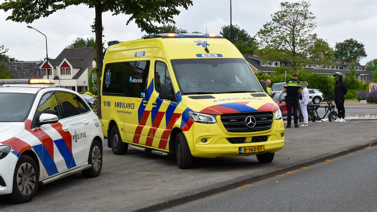 Een politieauto en een ambulance staan geparkeerd langs de weg. Op de achtergrond staan enkele politieagenten en mensen bij een fiets. Het weer is bewolkt.