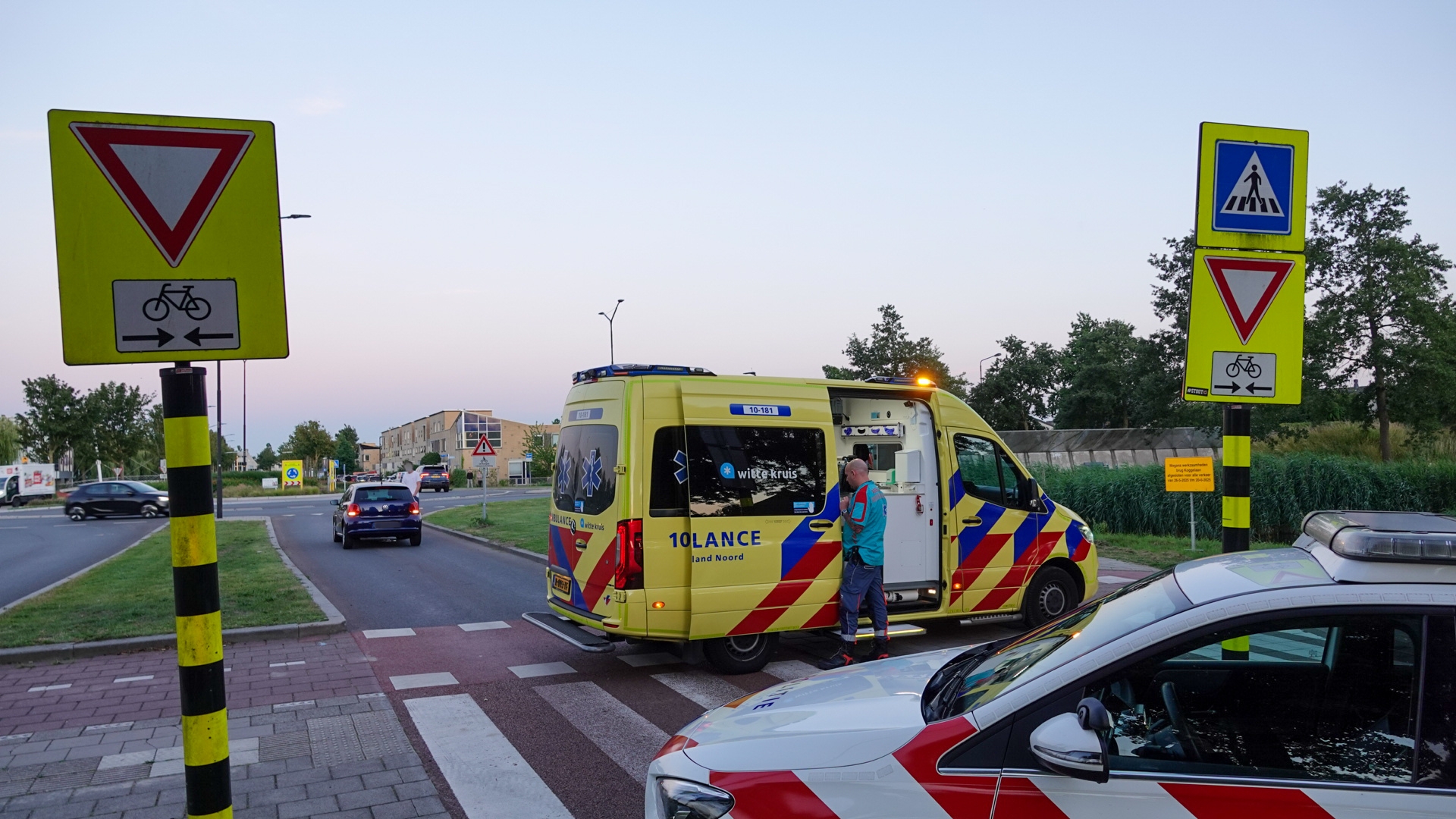 Een ambulance en een politieauto staan geparkeerd op een kruispunt bij een zebra- en fietspad, met een verkeersbord dat voorrang aangeeft.