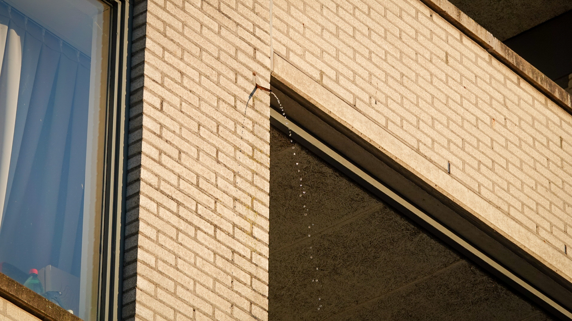 Buitenkant van een bakstenen gebouw met een afgeknikte waterafvoer waaruit druppels water vallen.