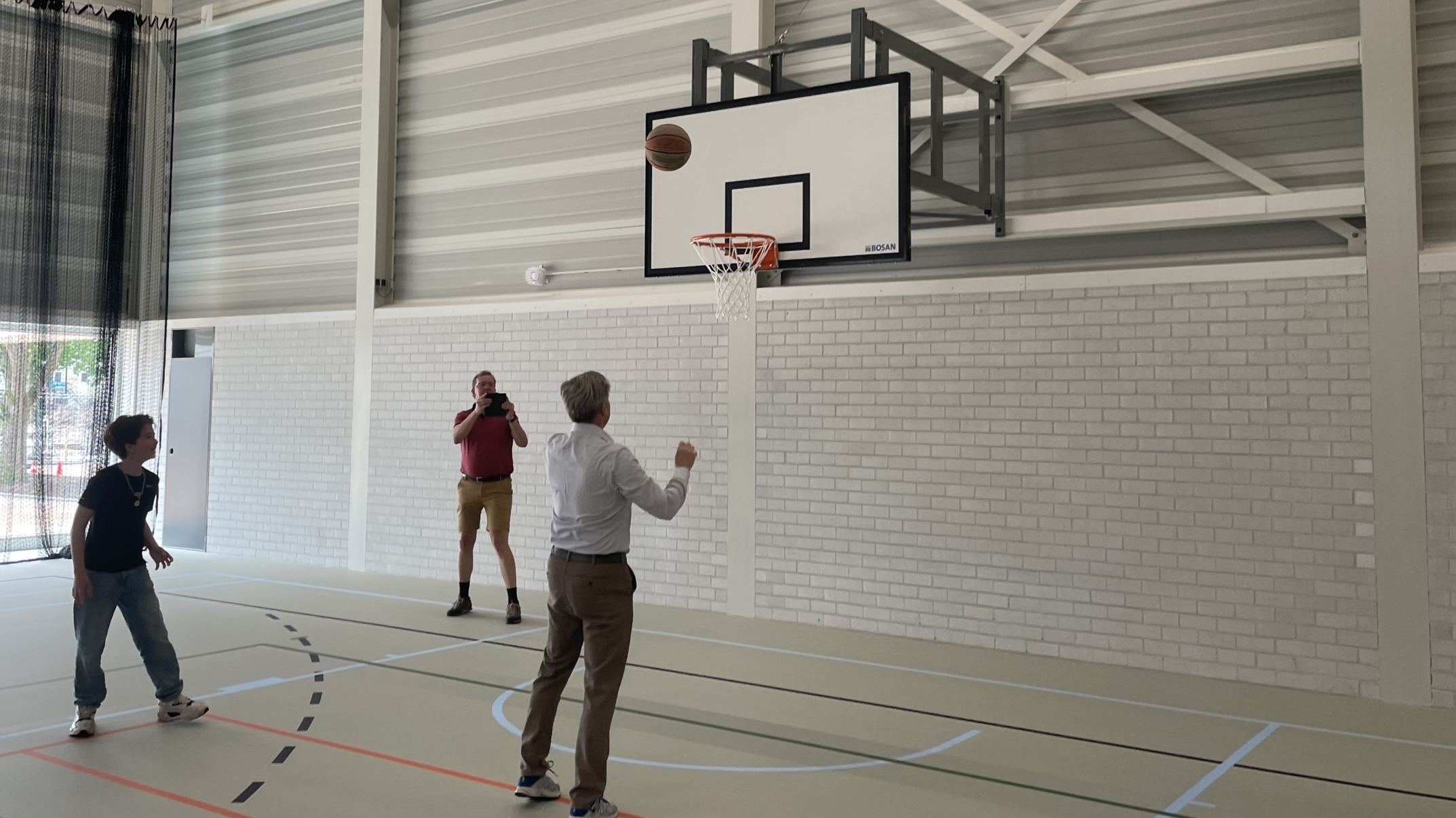 Een man in een lichte blouse gooit een basketbal naar een basket in een sporthal, terwijl een jongen en een andere man toekijken. Er zijn sportlijnen op de vloer en twee muren bestaan uit witte bakstenen en metalen panelen.