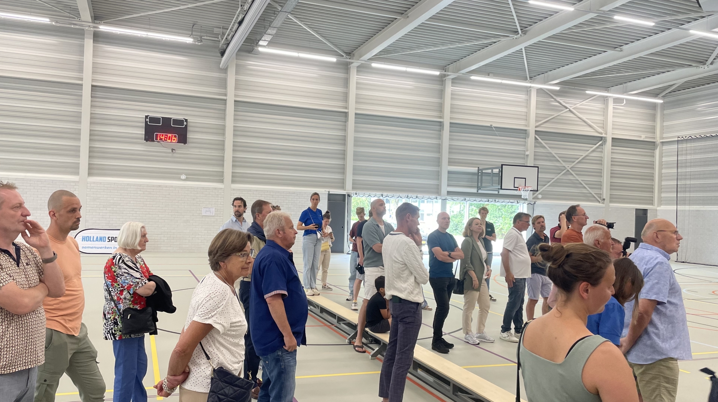 Groep mensen in een sporthal, sommigen staan op tribunes terwijl anderen rondlopen, met een basketbalnet en klok op de achtergrond.