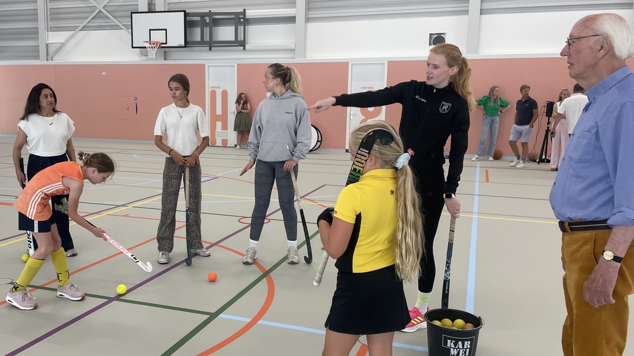 Kinderen en volwassenen in een gymzaal met hockey-uitrusting, waar een jonge speler klaar staat om een bal te slaan.