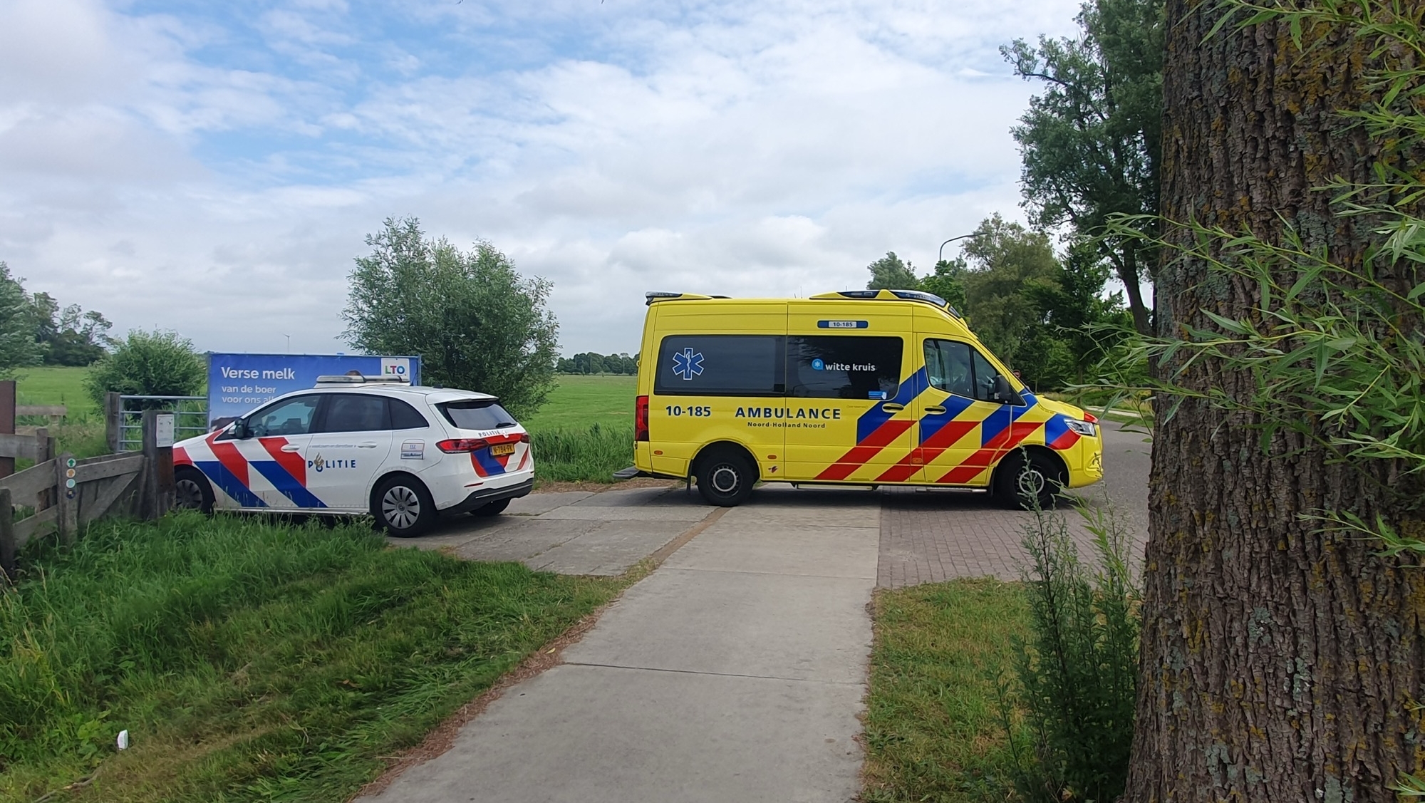 Een Nederlandse politieauto en ambulance staan geparkeerd naast een weiland aan de rand van een verharde weg.