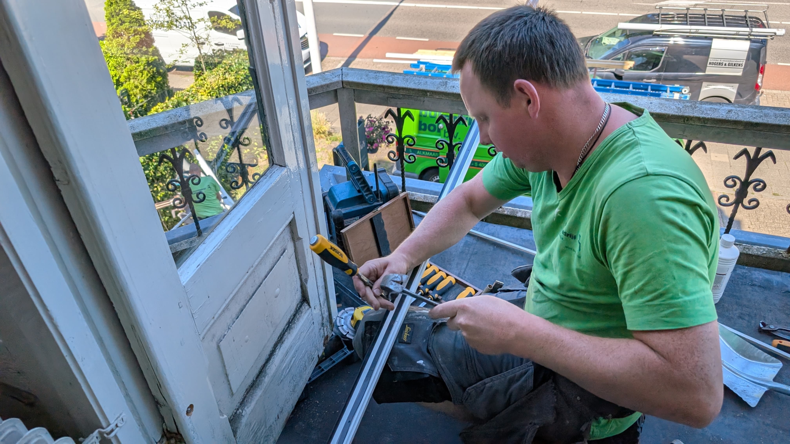 Een man in een groene shirt is bezig met gereedschap op een balkon, vermoedelijk met kluswerkzaamheden aan een deur of raamkozijn.