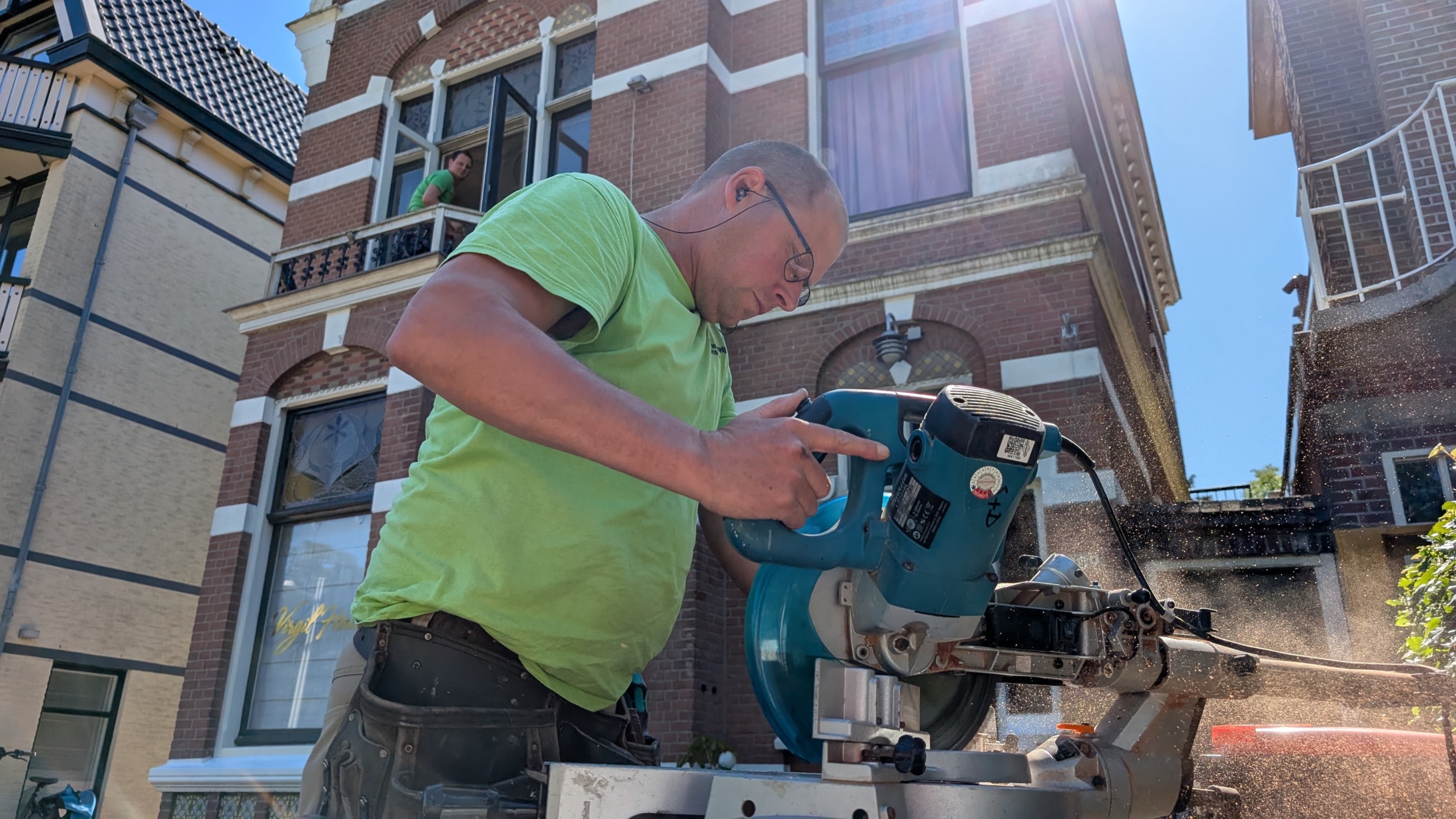 Een man met een koptelefoon zaagt met een elektrische zaag buiten een bakstenen gebouw, terwijl iemand vanuit een open raam toekijkt.