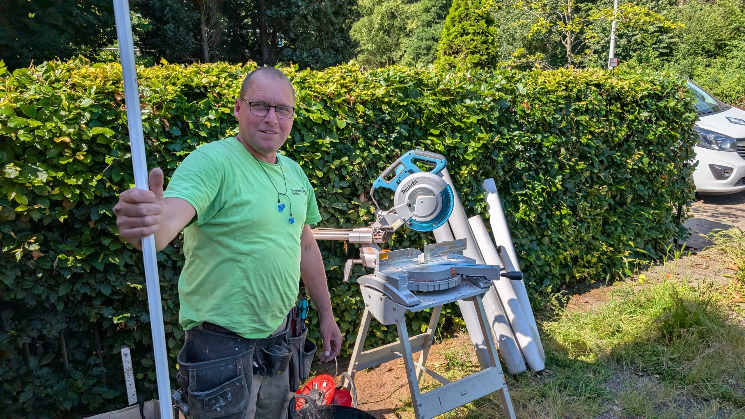 Man in a green shirt holding a long white pole next to a miter saw and standing in front of a hedge on a sunny day.