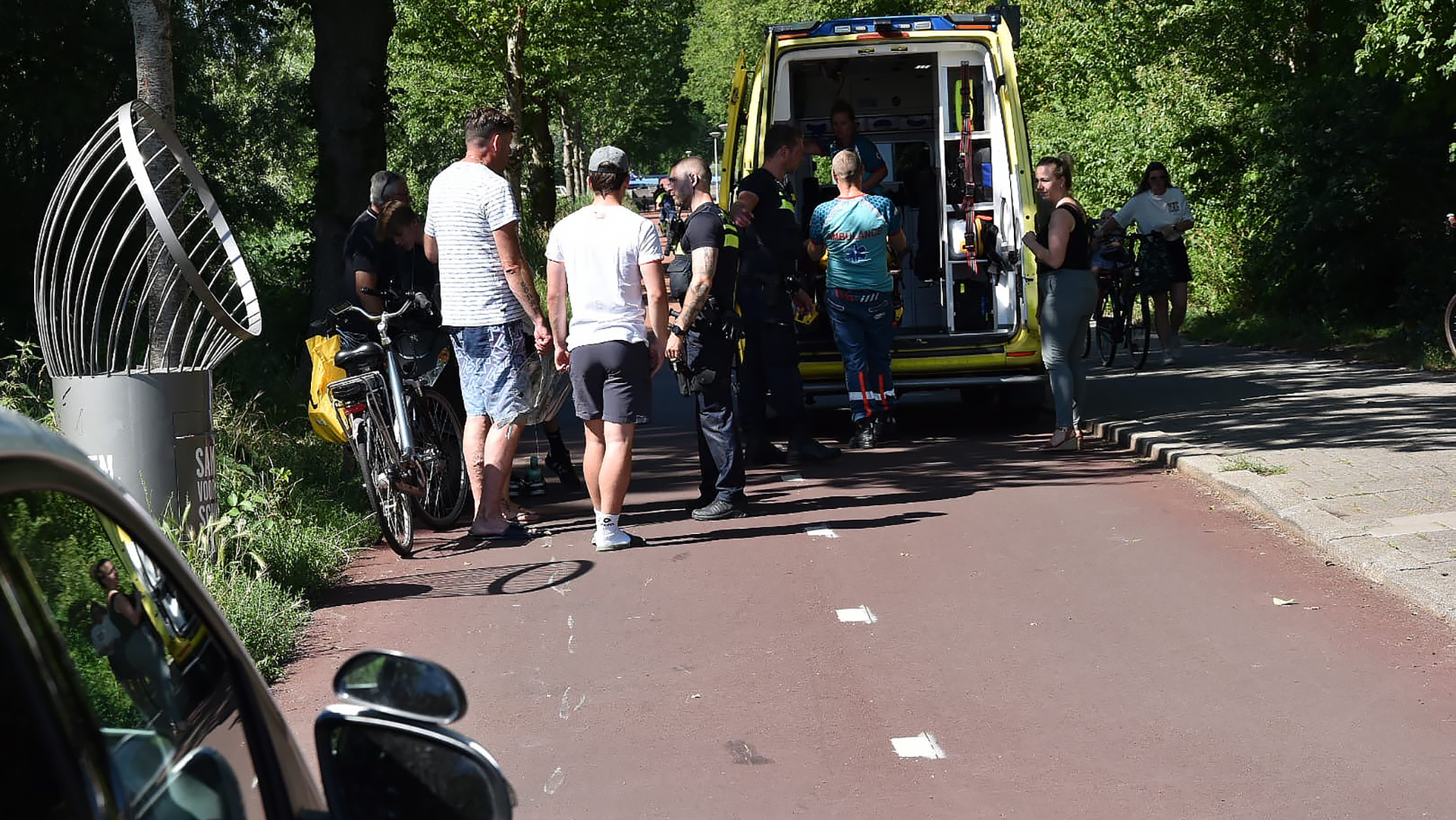 Groep mensen verzameld rond een ambulance op een fietspad in een groene omgeving; een paar fietsen zijn zichtbaar en paramedici behandelen iemand in het geopende voertuig.