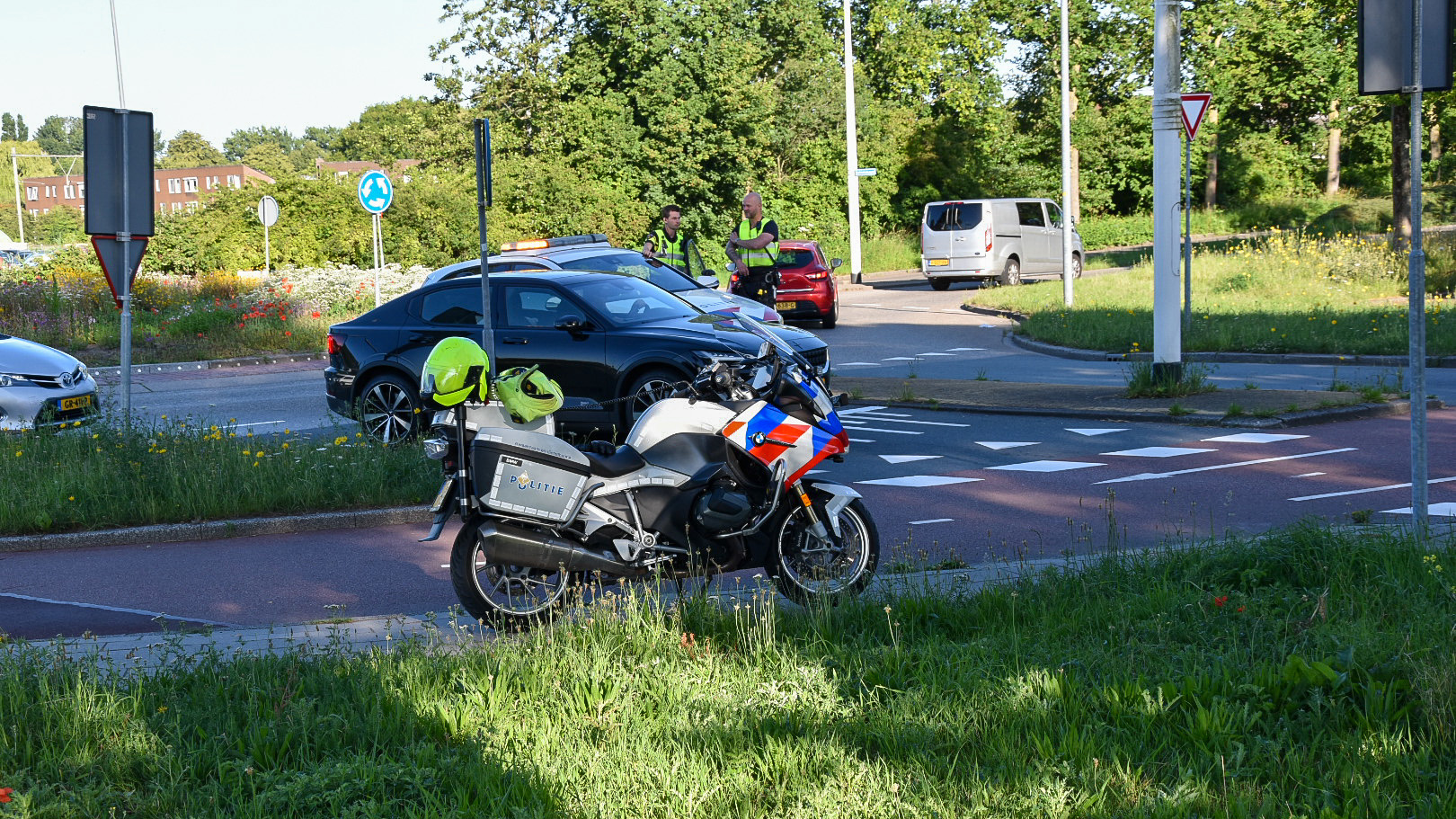 Een politie motorfiets stopt bij een rotonde met diverse voertuigen op de weg en twee politieagenten in reflecterende kleding die met elkaar praten. Op de achtergrond zijn groene struiken en bomen te zien.