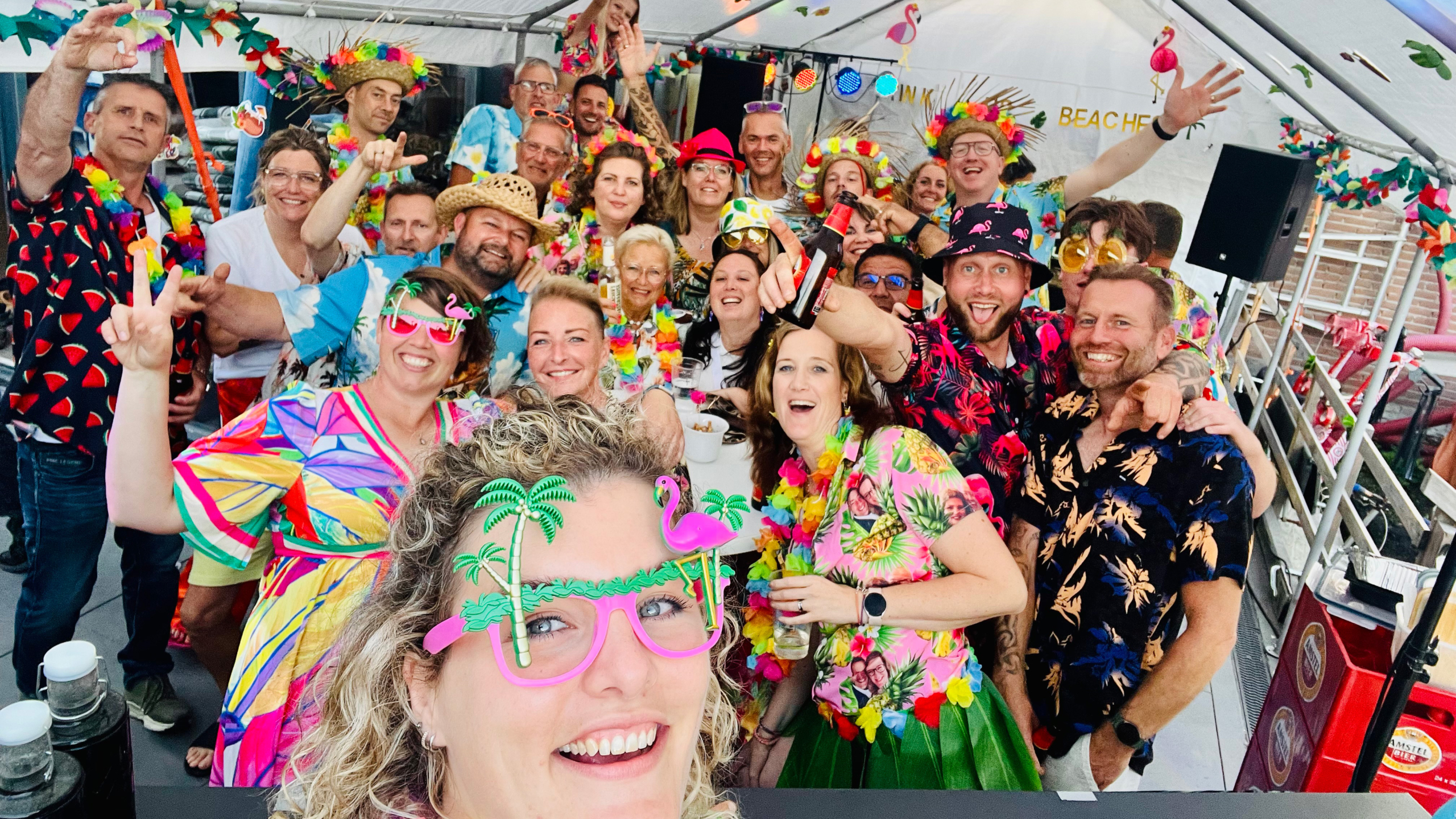 Een groep mensen in vrolijke, kleurrijke kleding en hawaiikrans poseren lachend onder een versierde tent.