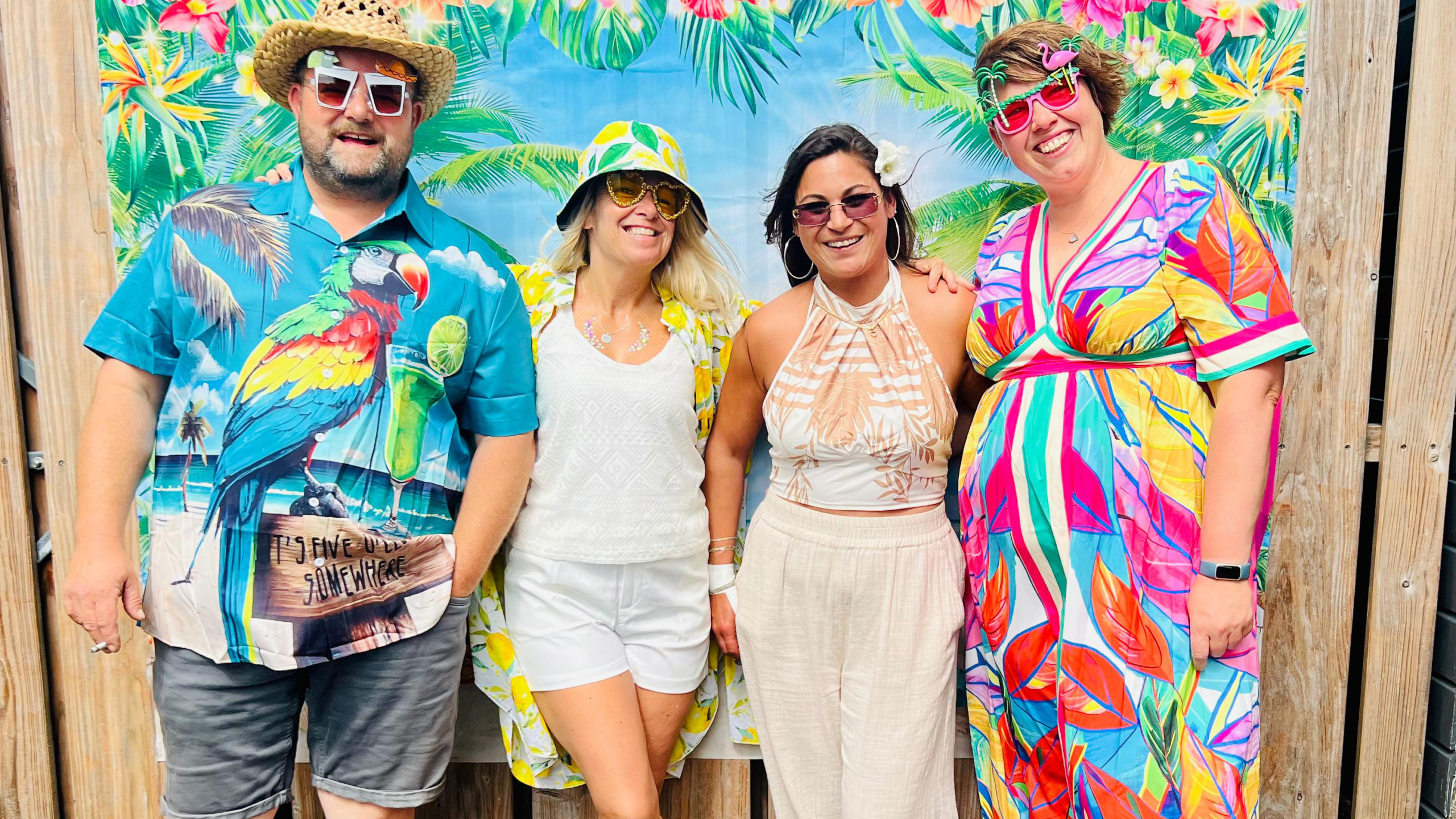 Vier mensen in kleurrijke zomerkleding staan samen voor een tropische achtergrond met palmbomen en bloemen. Ze dragen zonnebrillen en glimlachen naar de camera. De achtergrond en outfits geven een vrolijke en zomerse sfeer weer.