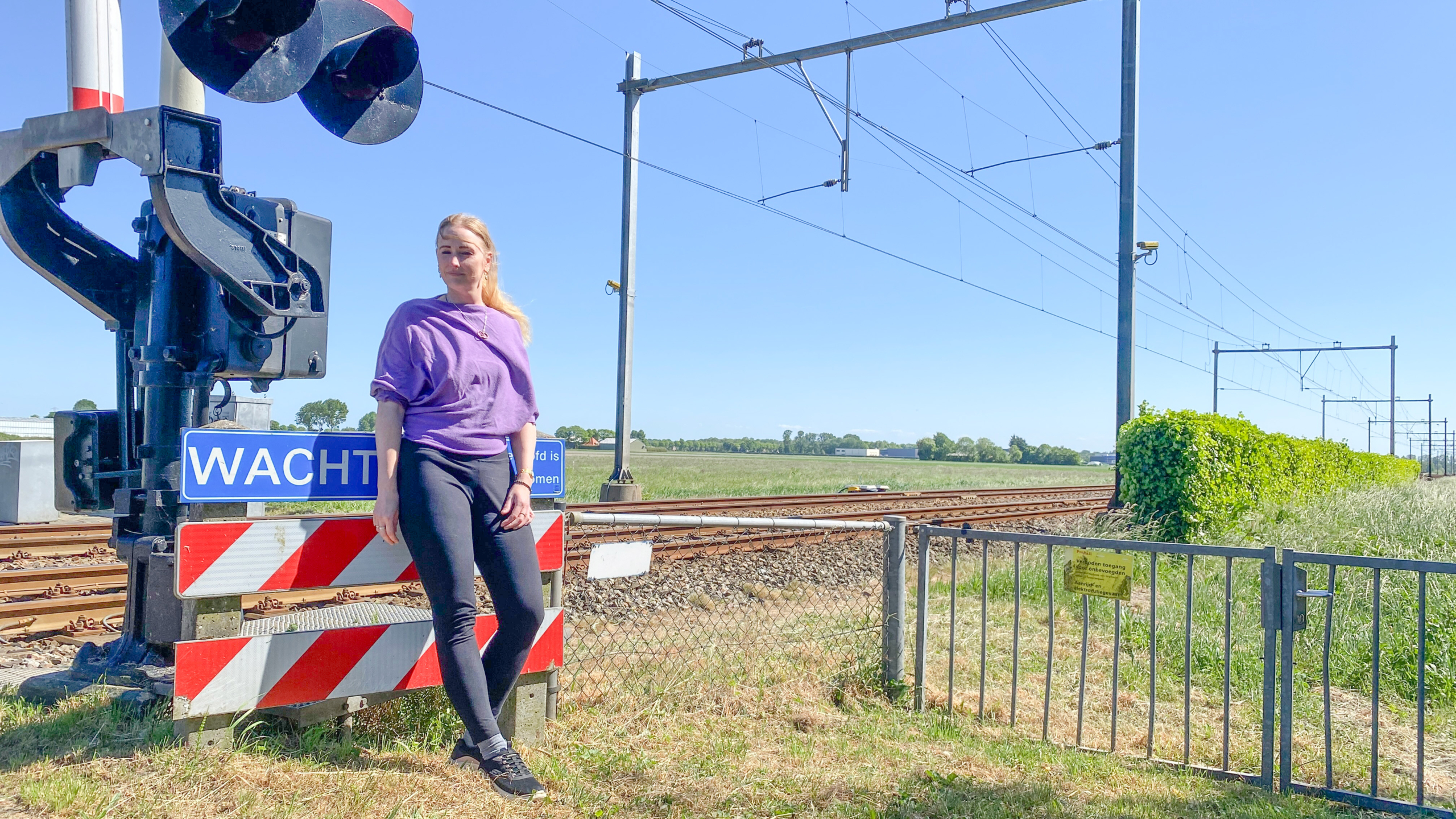Een vrouw in een paarse trui en zwarte broek staat naast een spoorweg overgang met een "WACHT" bord, onder een heldere blauwe lucht.