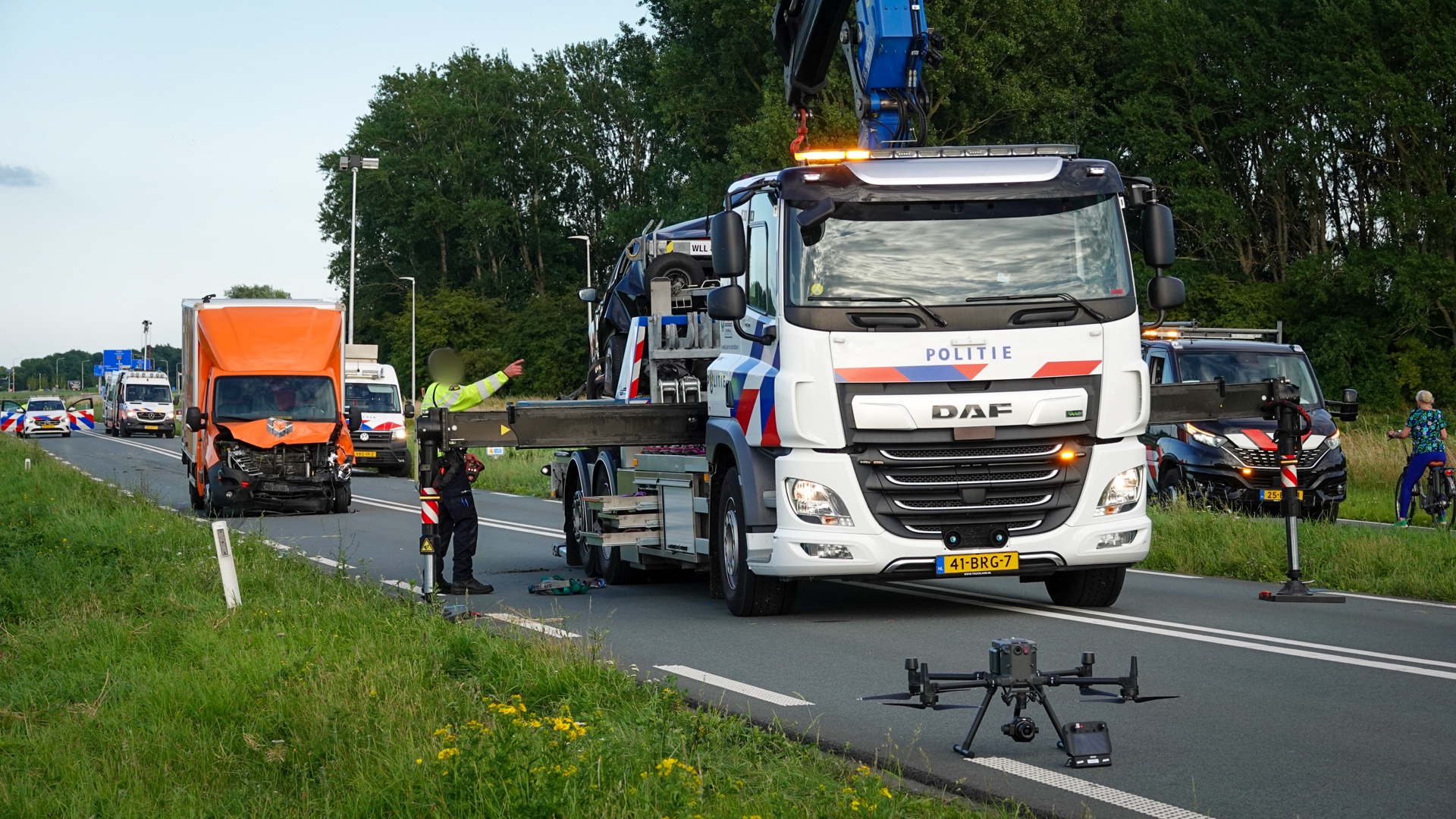 Vrachtwagens en bestelwagens staan op een weg met politievoertuigen; een bergingsvoertuig is bezig met het opruimen van voertuigen, terwijl een grote drone op de voorgrond op het wegdek staat.
