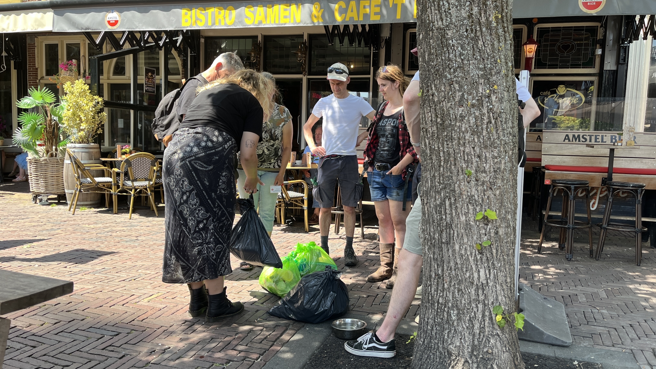 Mensen verzamelen zich rond zakken afval op een zonnige stoep voor een bistro en café genaamd 'Bistro Samen & Café'.