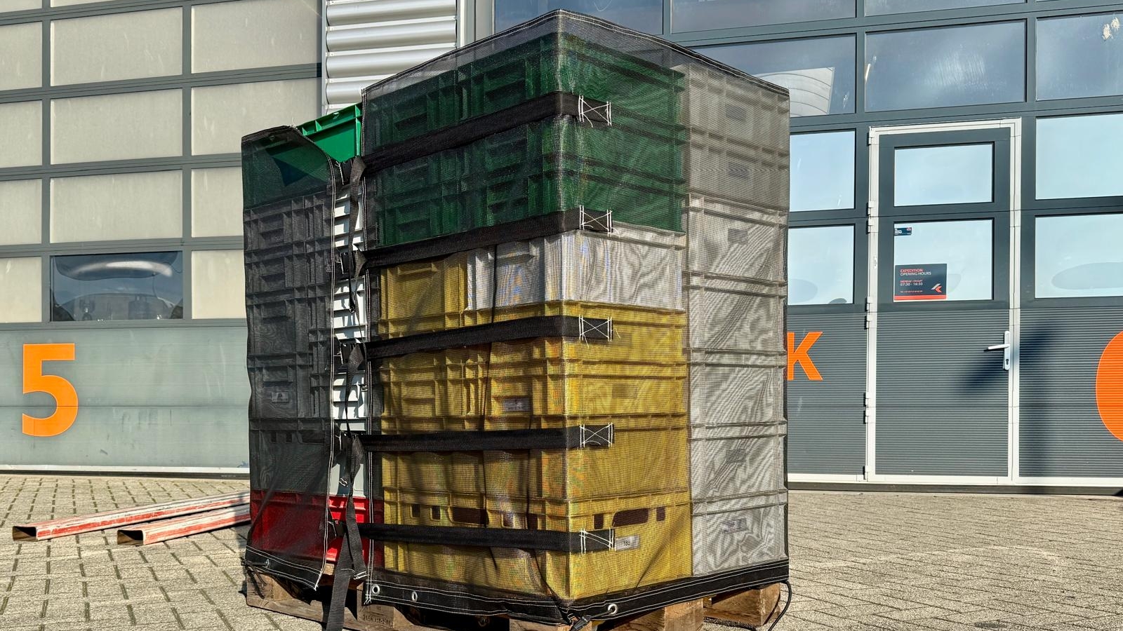 Een palet met gestapelde kratten bedekt met een net, tegen een industriële achtergrond met een groot nummer vijf en een deel van een K zichtbaar op een gebouw.