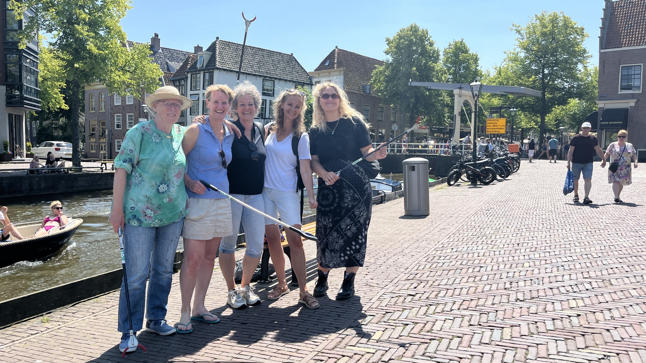Vijf mensen poseren op een zonnig terras aan een gracht, met grijpstokken in hun handen. Ze staan dichtbij elkaar en lachen naar de camera. Op de achtergrond is een brug, fietsen en langsvarende boot te zien.