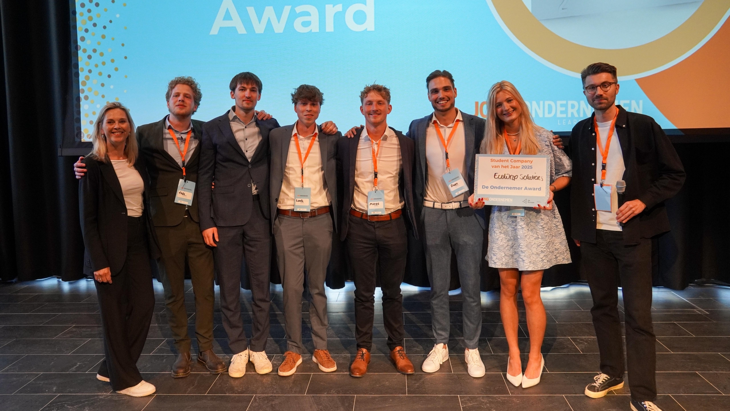 Groep mensen poseert op het podium met een certificaat voor de "De Ondernemer Award". Op de achtergrond is een scherm met de tekst "De Ondernemer Award". Alle personen dragen naamkaartjes om hun nek en lachen in de camera.
