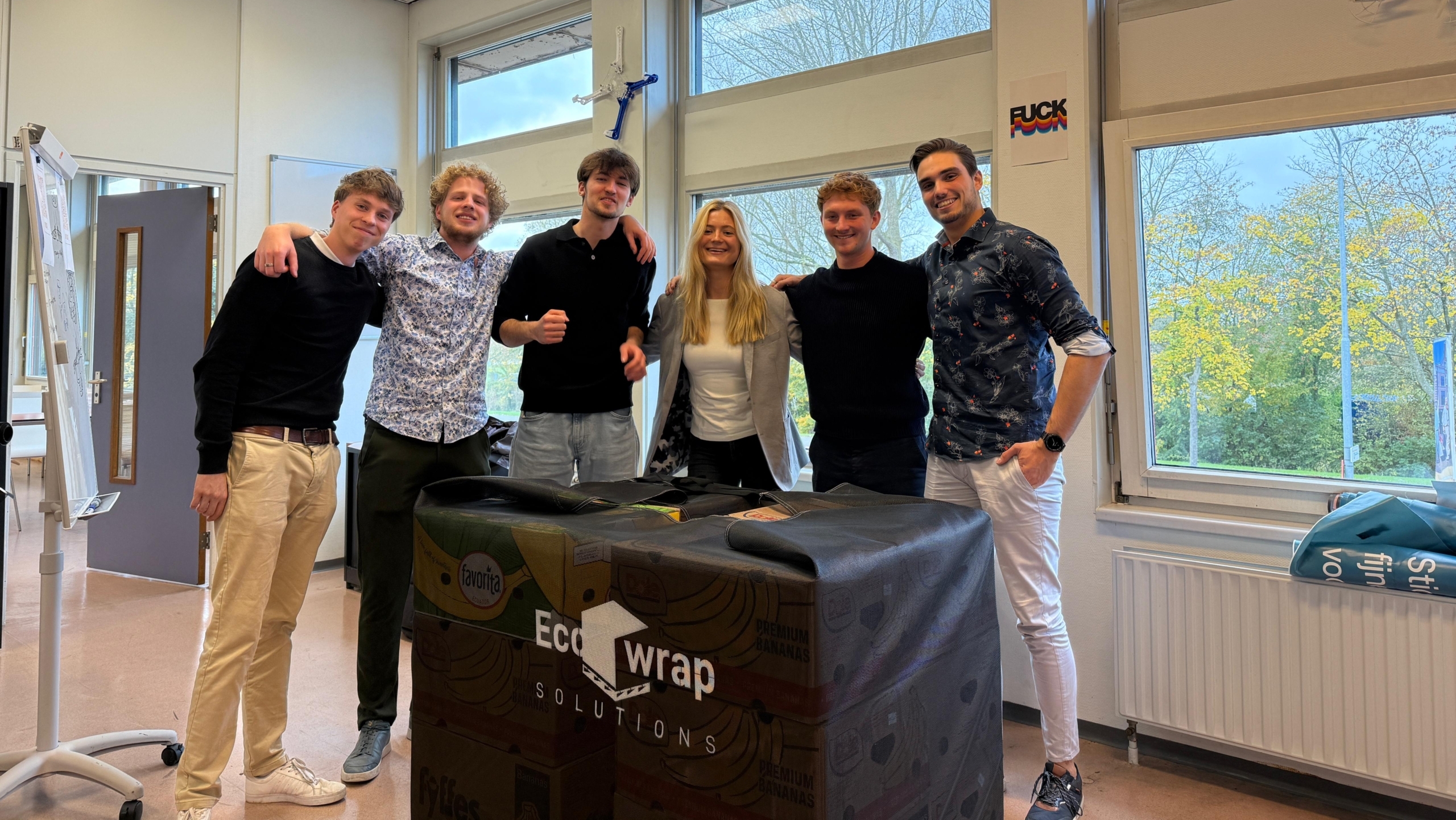 Een groep van zes mensen poseren samen in een klaslokaal met grote ramen en een flipover. De voorkant van het klaslokaal is bedekt met een groot Eco Wrap Solutions doek. Buiten de ramen zijn bomen met herfstkleuren zichtbaar. De sfeer is informeel en vriendschappelijk.