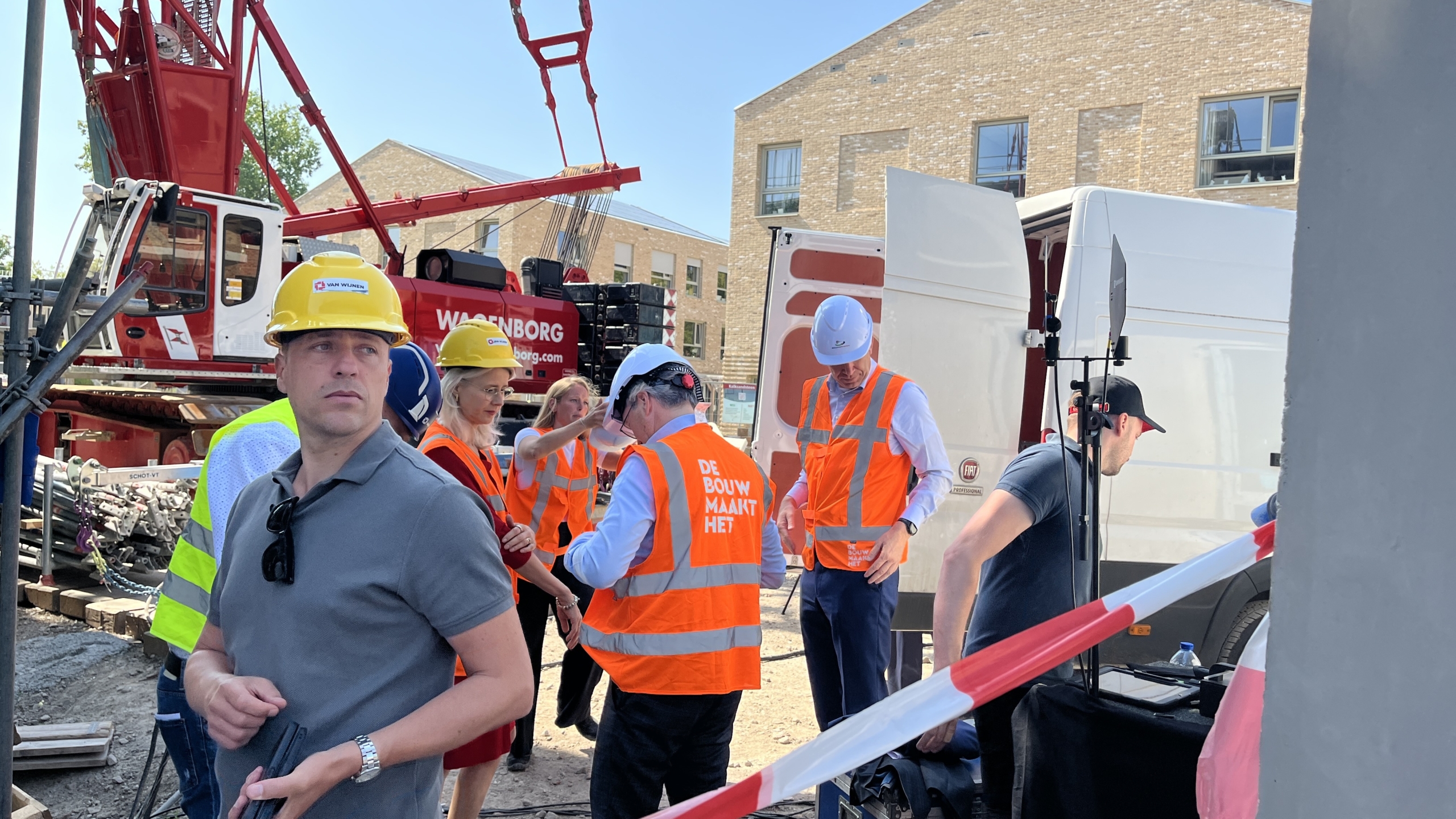 Mensen in veiligheidskleding, waaronder helmen en oranje hesjes, staan bij een bouwplaats met een kraan en een witte bestelwagen op de achtergrond.
