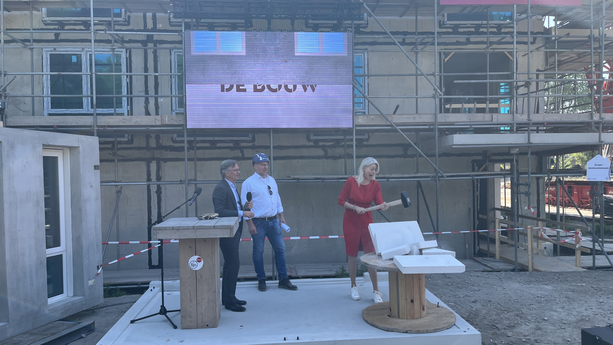 Een vrouw in een rode jurk houdt een hamer vast naast twee mannen in pakken voor een bouwplaats met een groot scherm op de achtergrond, waarop "DE BOUW" staat.