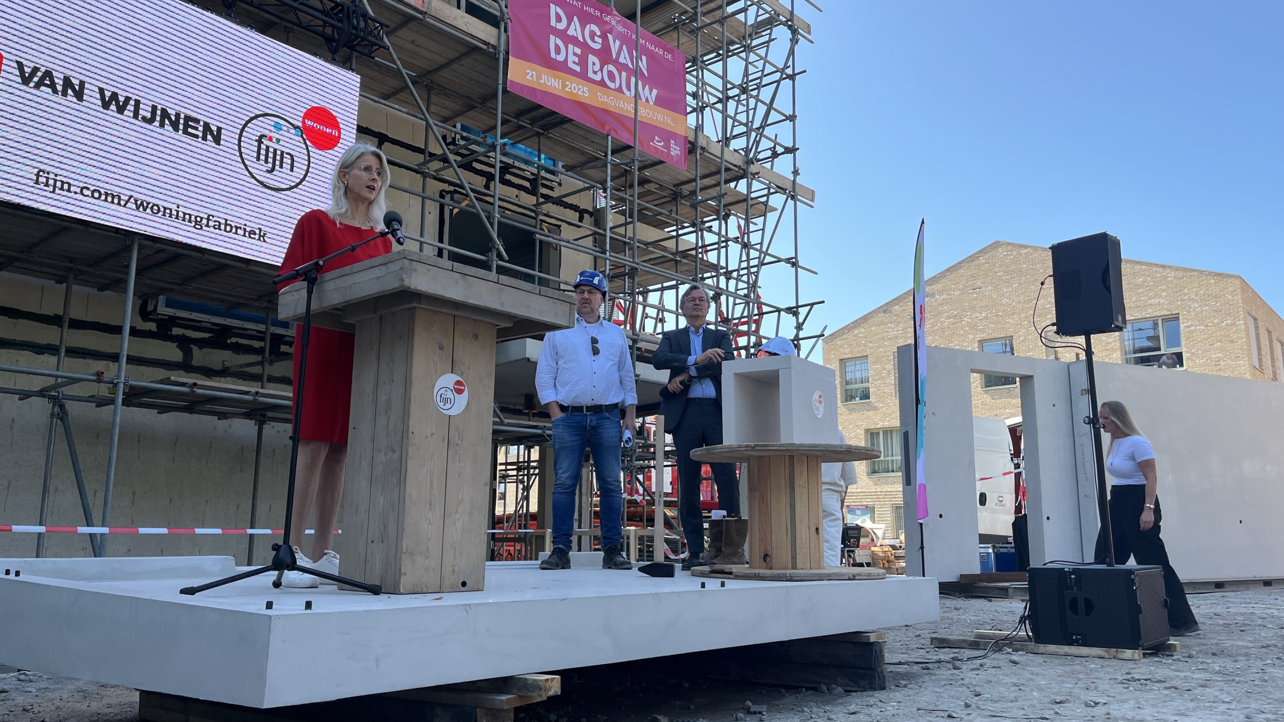 Een vrouw in een rode jurk spreekt op een podium tijdens een bouw evenement terwijl twee mannen naast haar staan; op de achtergrond is een banner met "Dag van de Bouw" te zien boven een steiger.