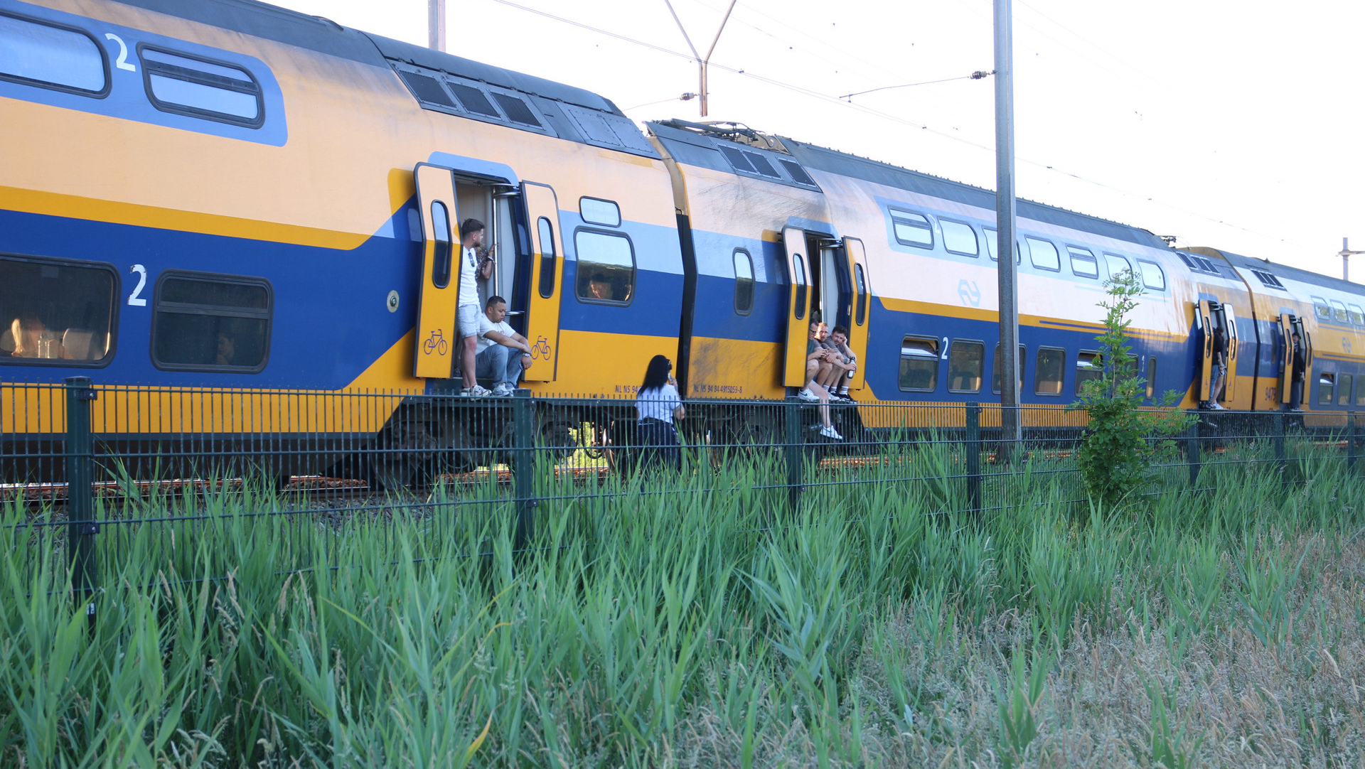 Een stilstaande dubbeldekstrein van de NS met geopende deuren waar mensen in en uit stappen, naast een grasveld en hekwerk.